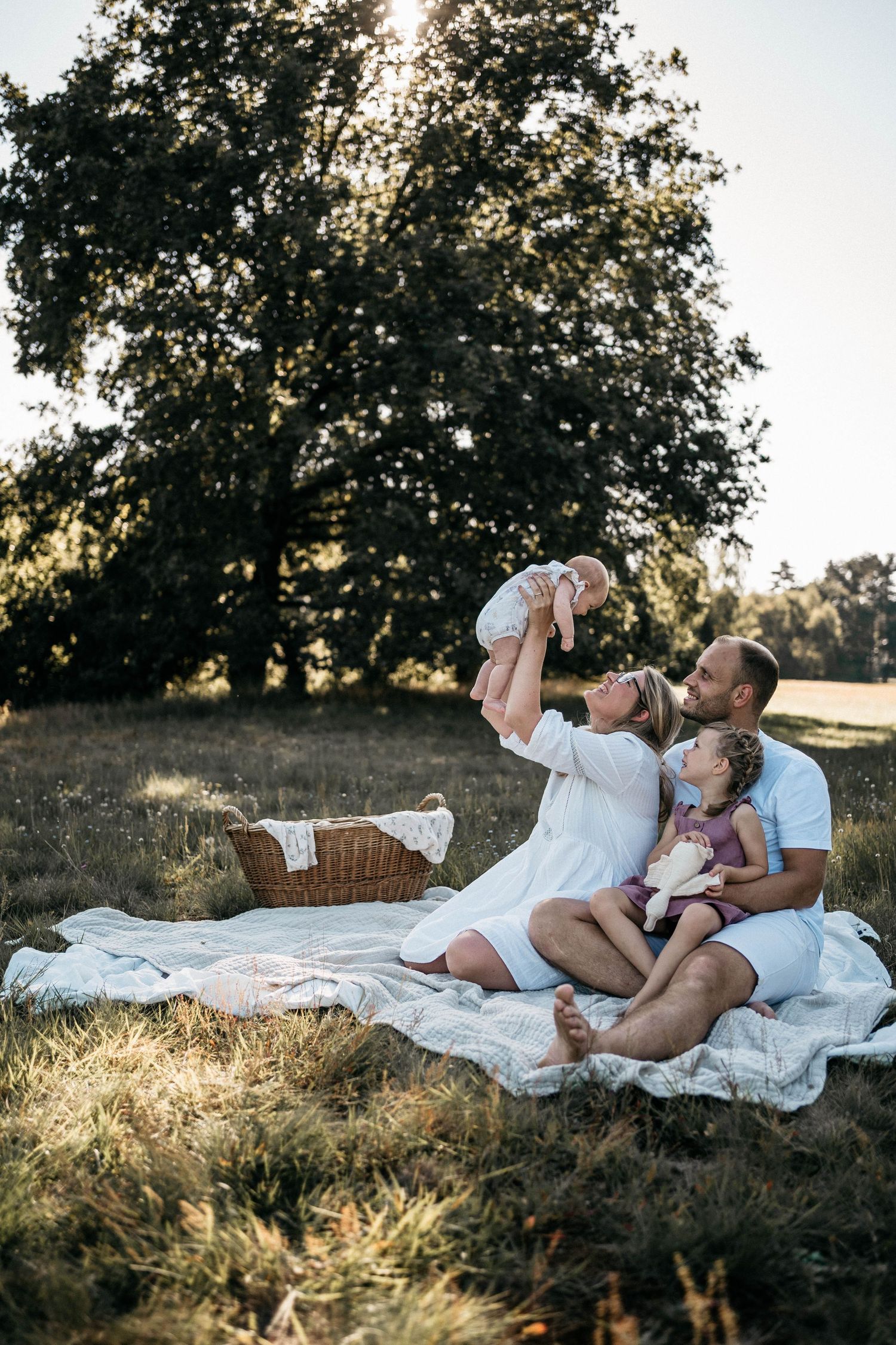Family picnic on white blanket under large tree during golden hour sunset.