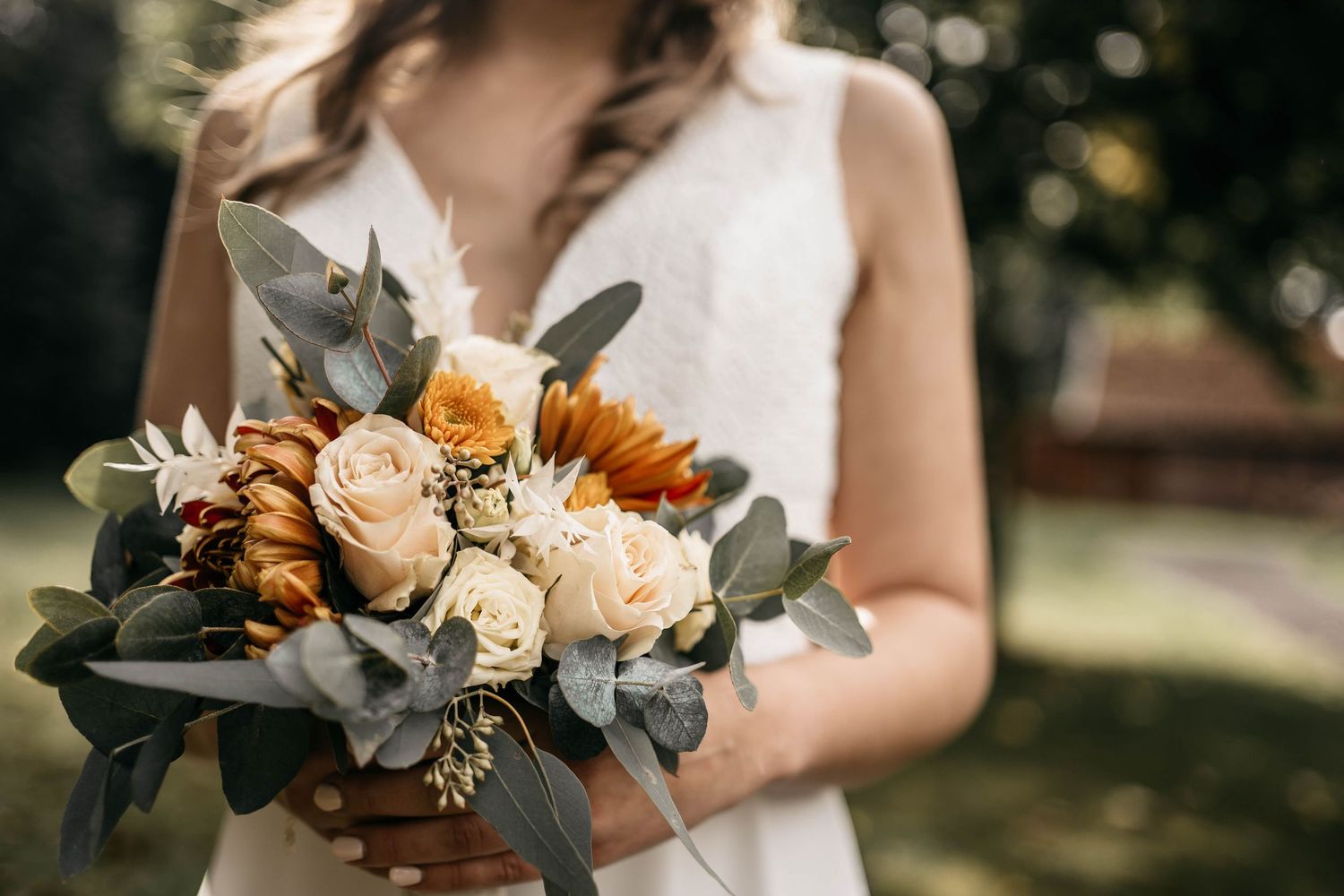 A wedding bouquet with orange and cream roses mixed with eucalyptus leaves held against a white dress in sunlit garden.