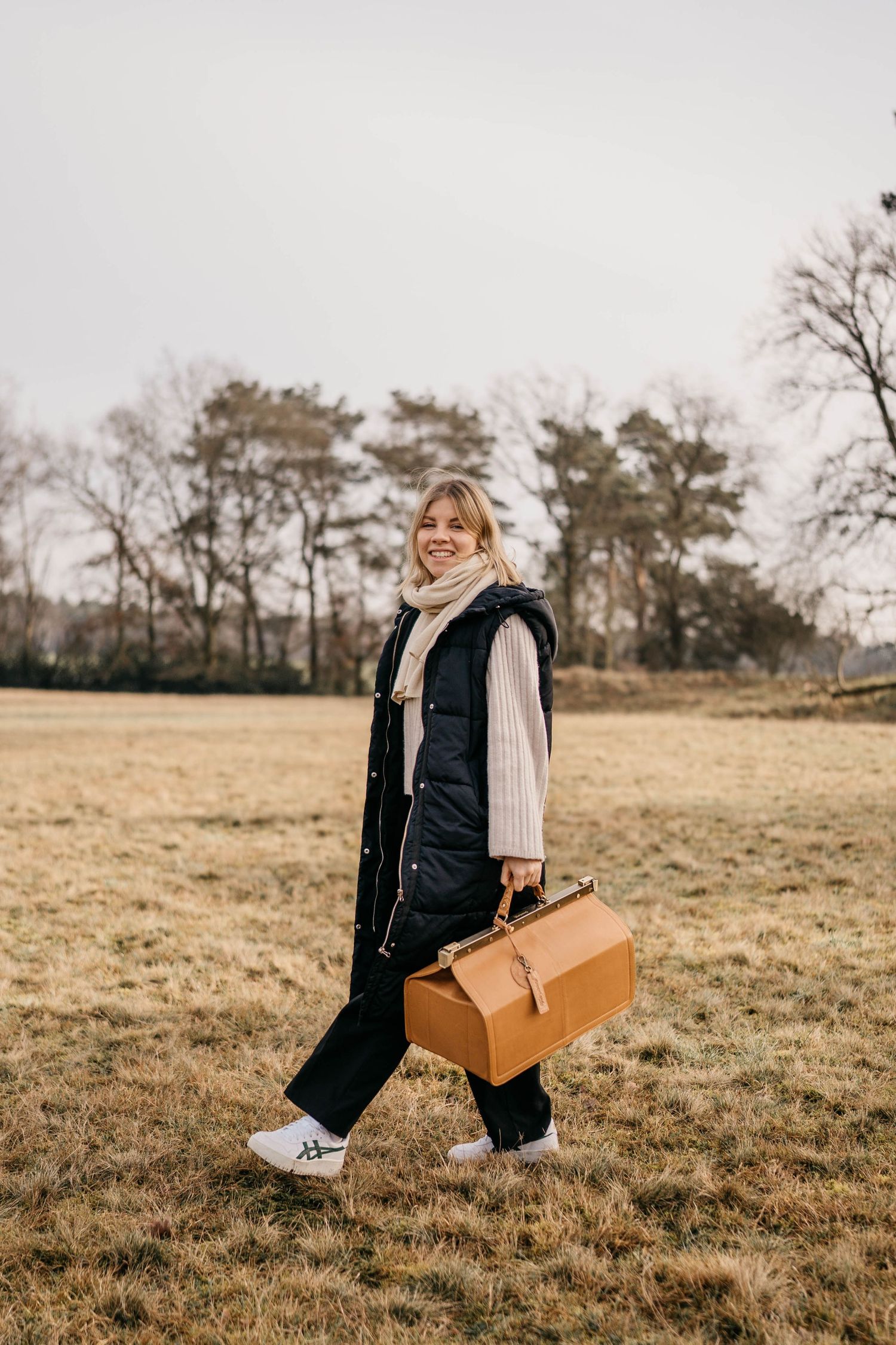 Person walking in a field wearing a black puffer vest, cream sweater and carrying a tan leather bag on an overcast day.
