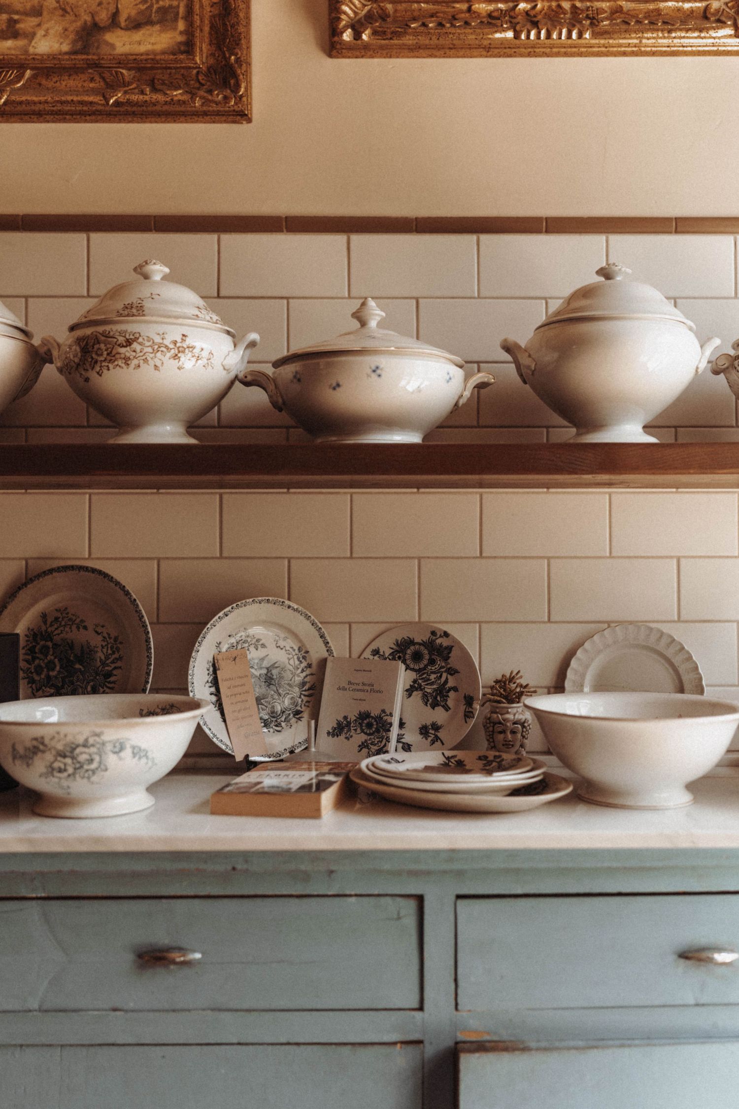 Vintage white and blue china dishes including tureens and plates displayed on wooden shelves above a blue-green cabinet.