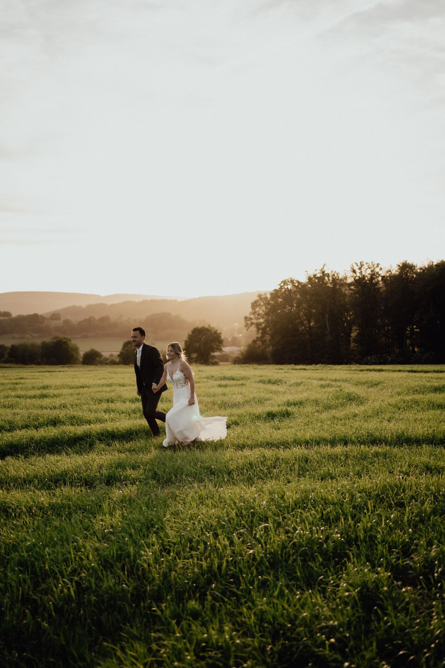 A couple walks together in a sunlit grassy meadow at golden hour with trees and rolling hills in the background.