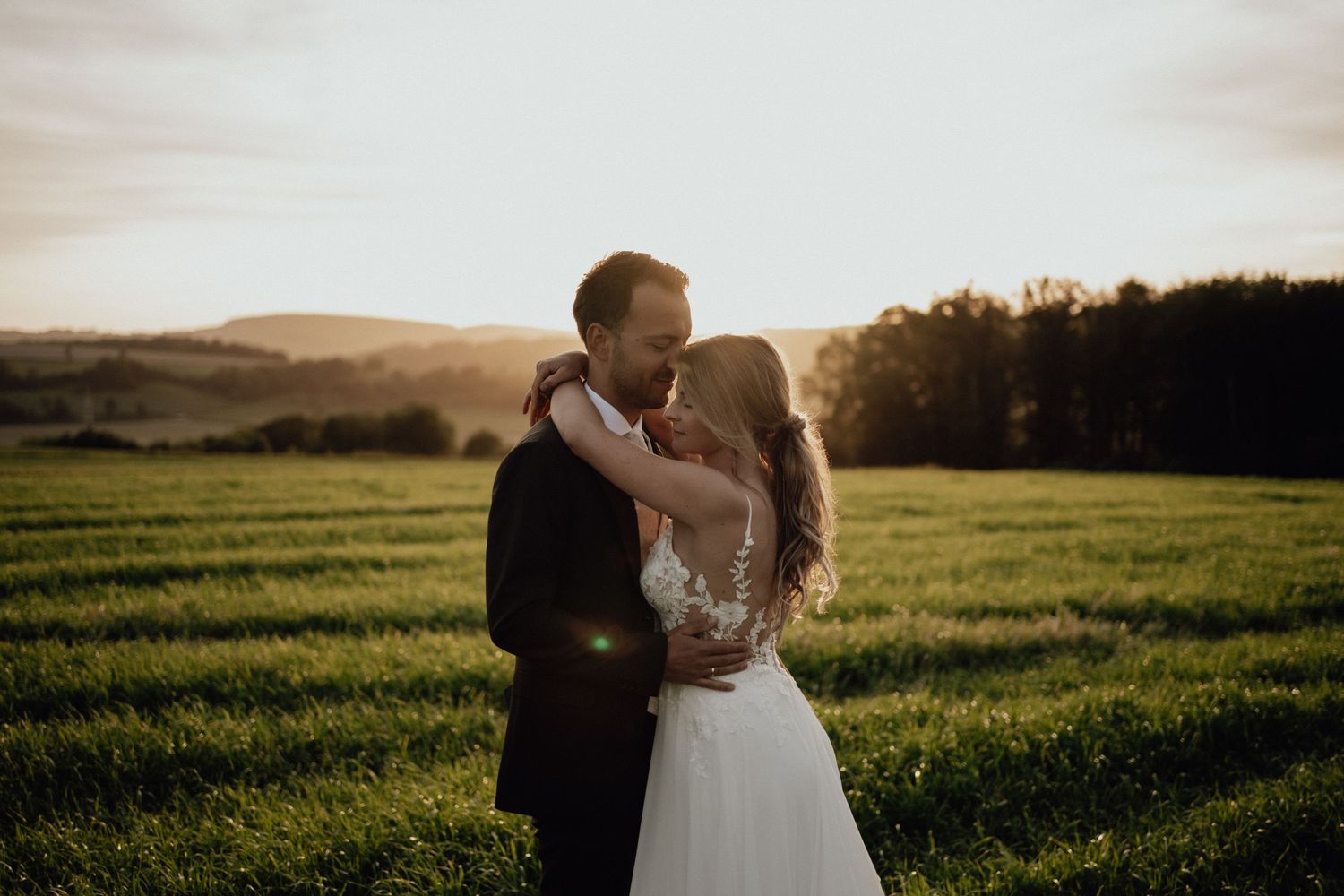 A couple embraces in a romantic sunset moment on a grassy hillside with golden light illuminating the scenic countryside.