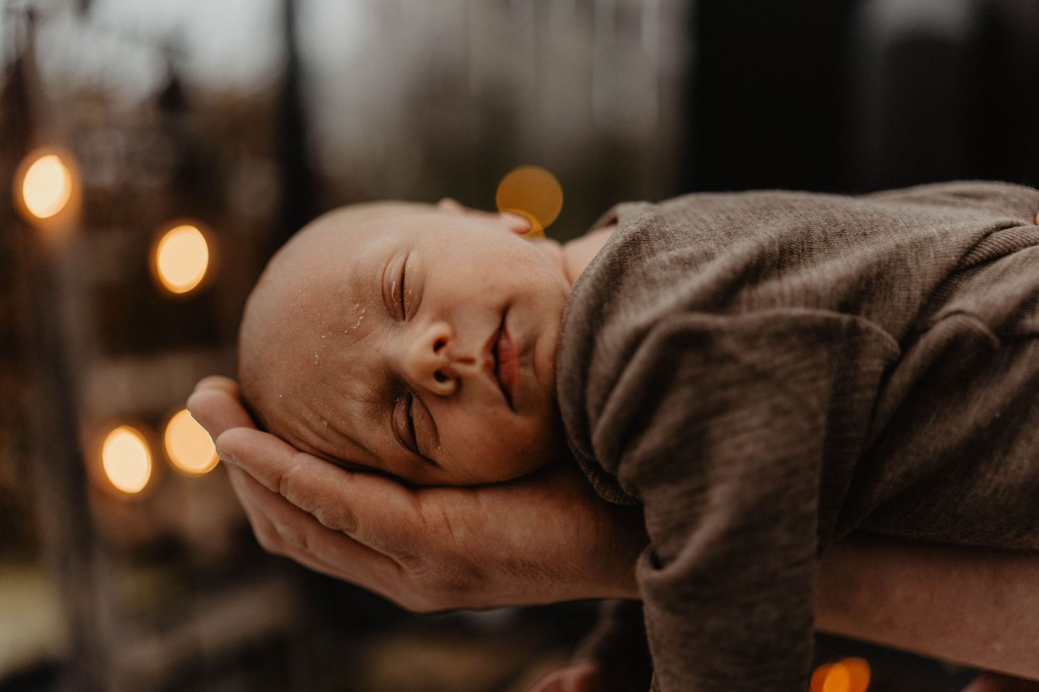 Sleeping baby in gray outfit with warm bokeh lights in background.