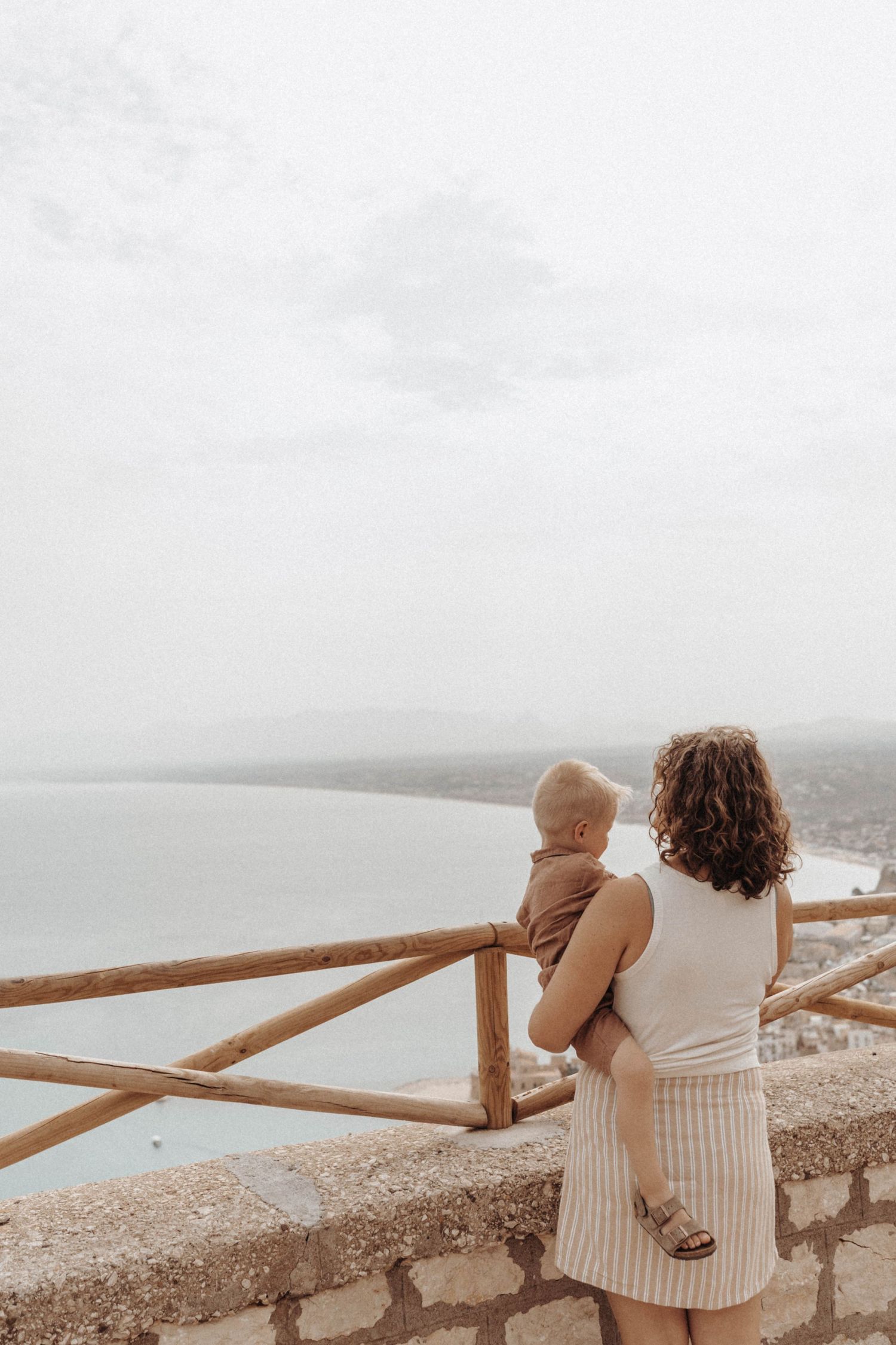 A person in a white knit dress holds a baby while looking out over a misty coastal view from a stone wall overlook.