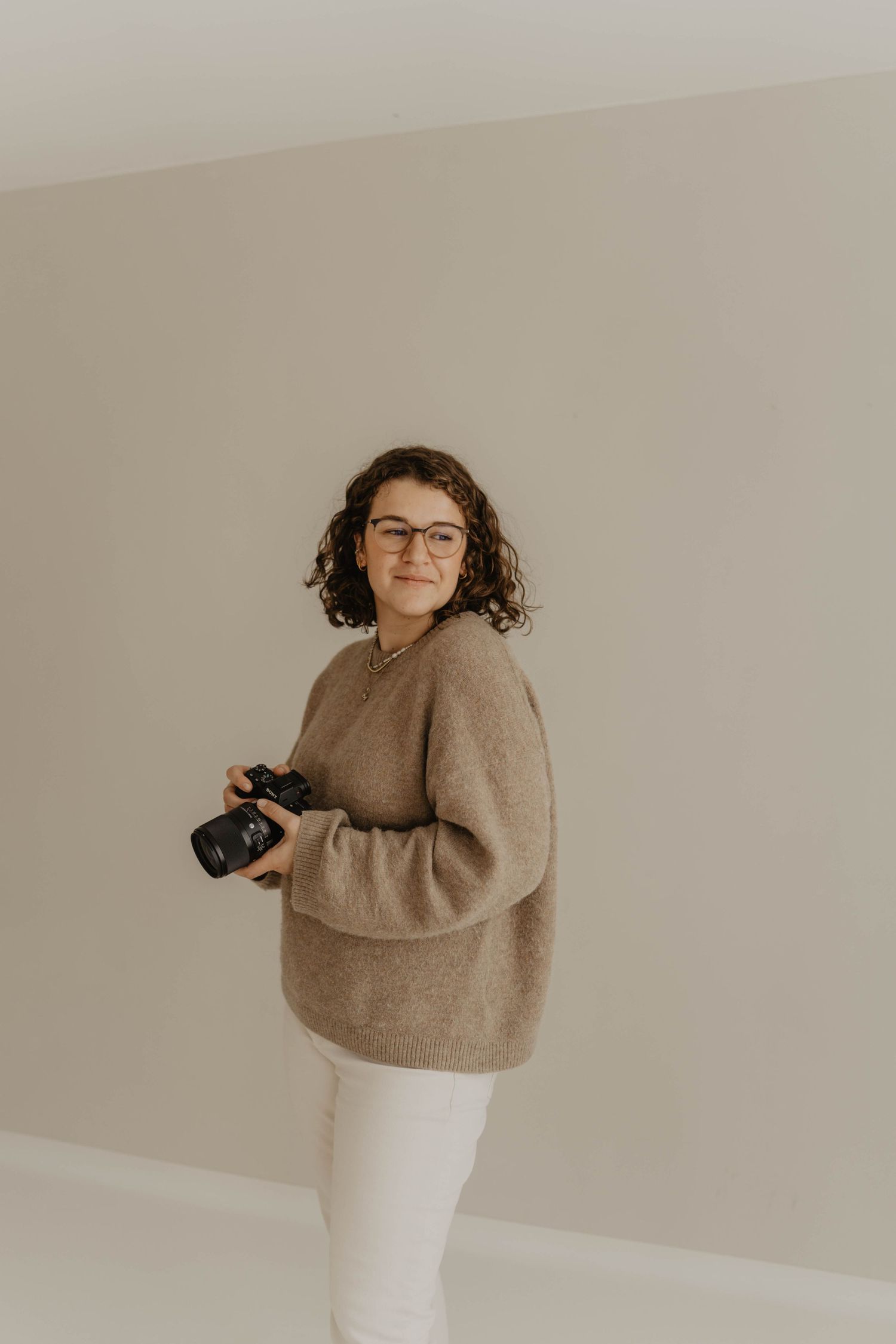 Woman in beige sweater holding camera against neutral wall background.