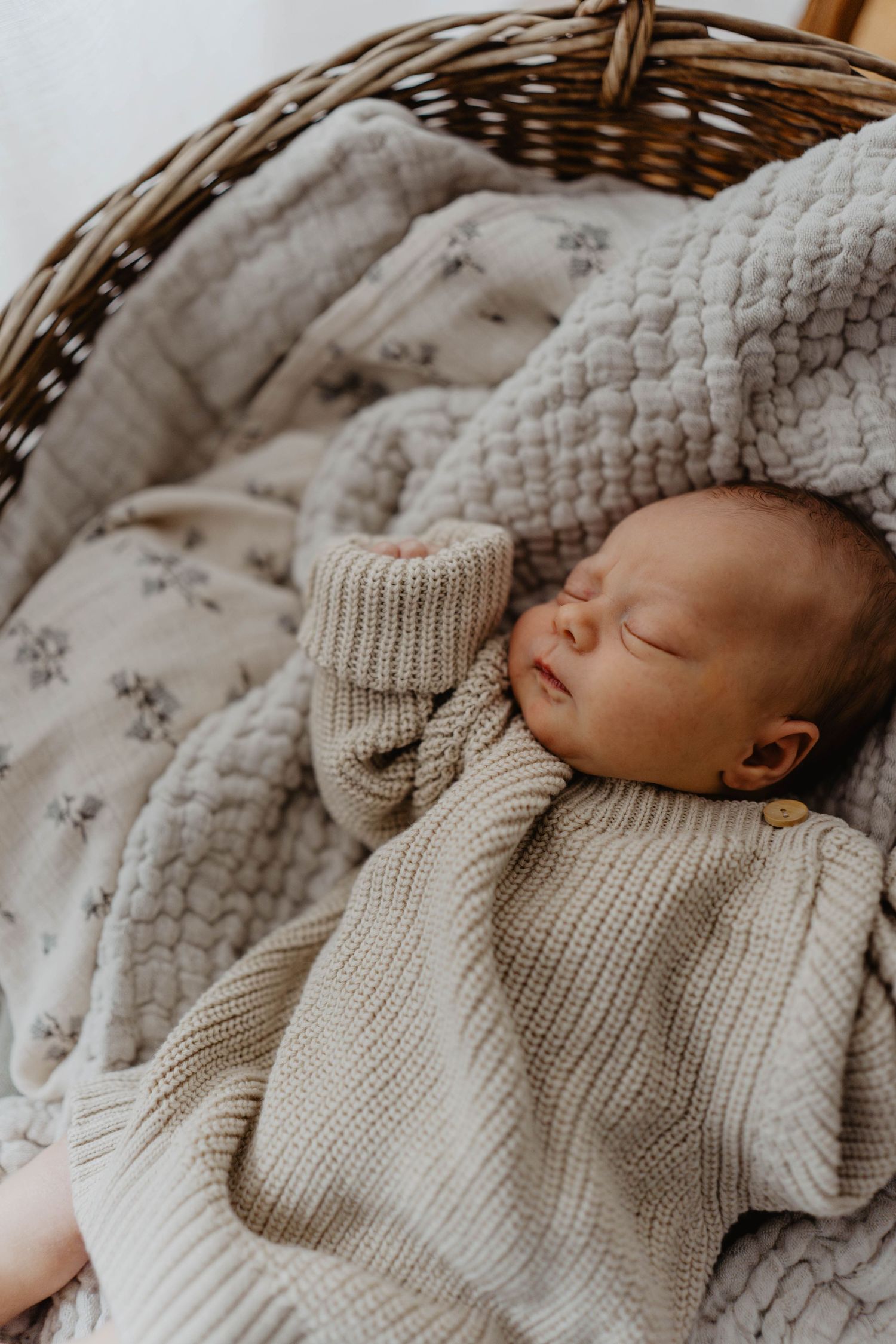Sleeping newborn in knit sweater resting peacefully in woven basket with textured blankets.