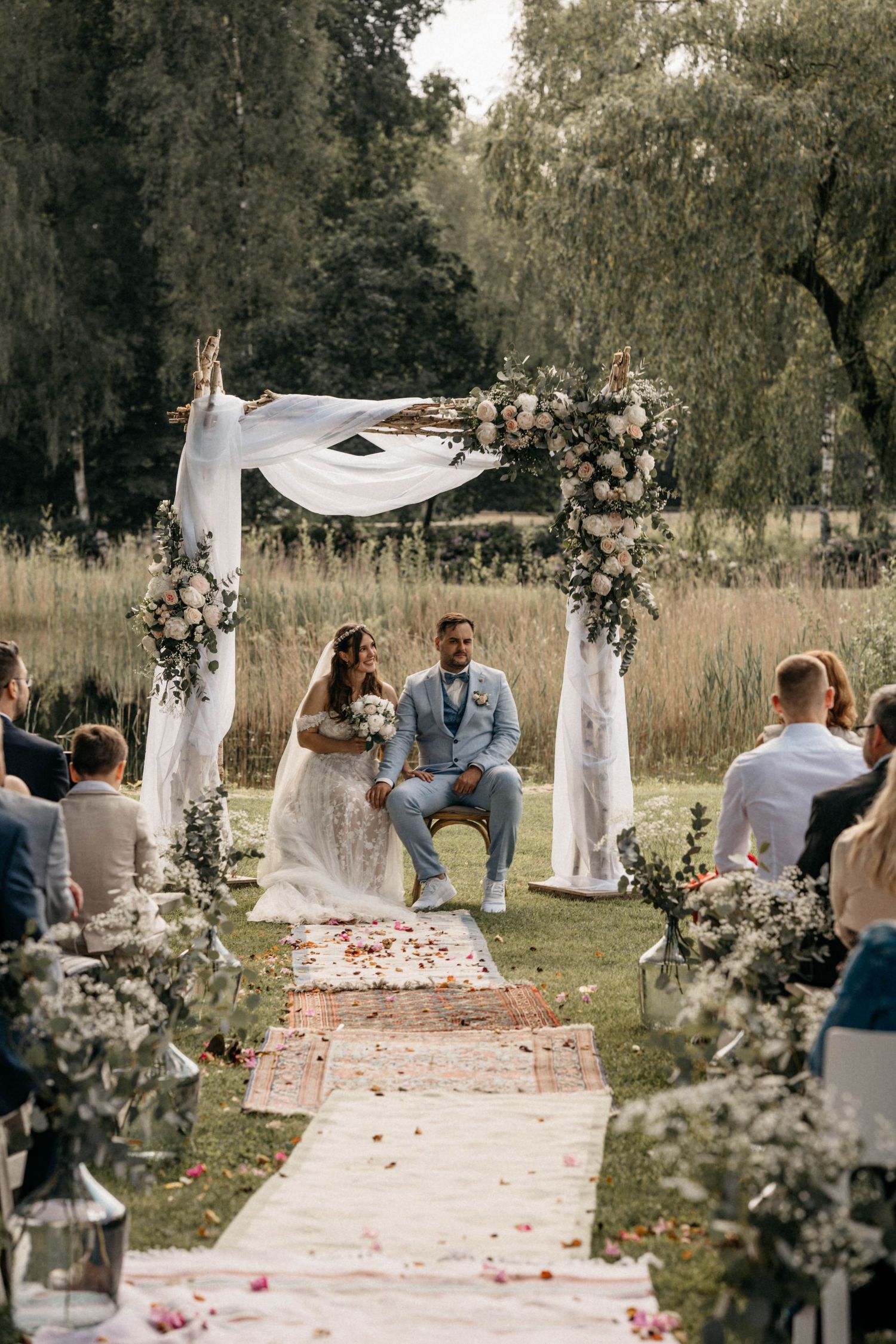 A romantic outdoor wedding ceremony setup with white draped arch and floral decorations along a rustic aisle runner in nature.