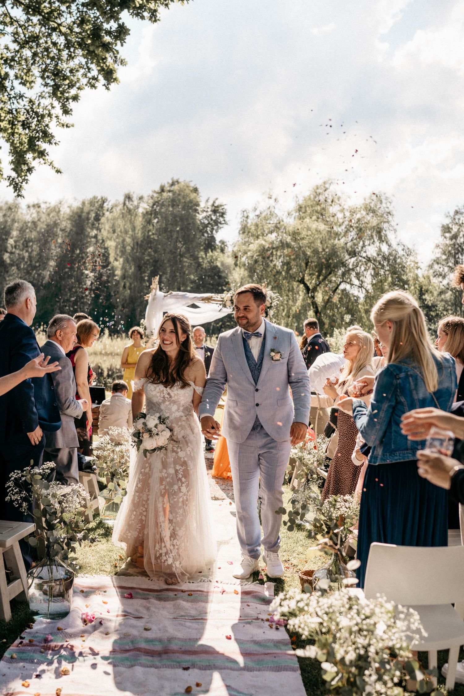 Newlyweds walk down outdoor aisle while guests toss flower petals during sunny garden wedding ceremony.