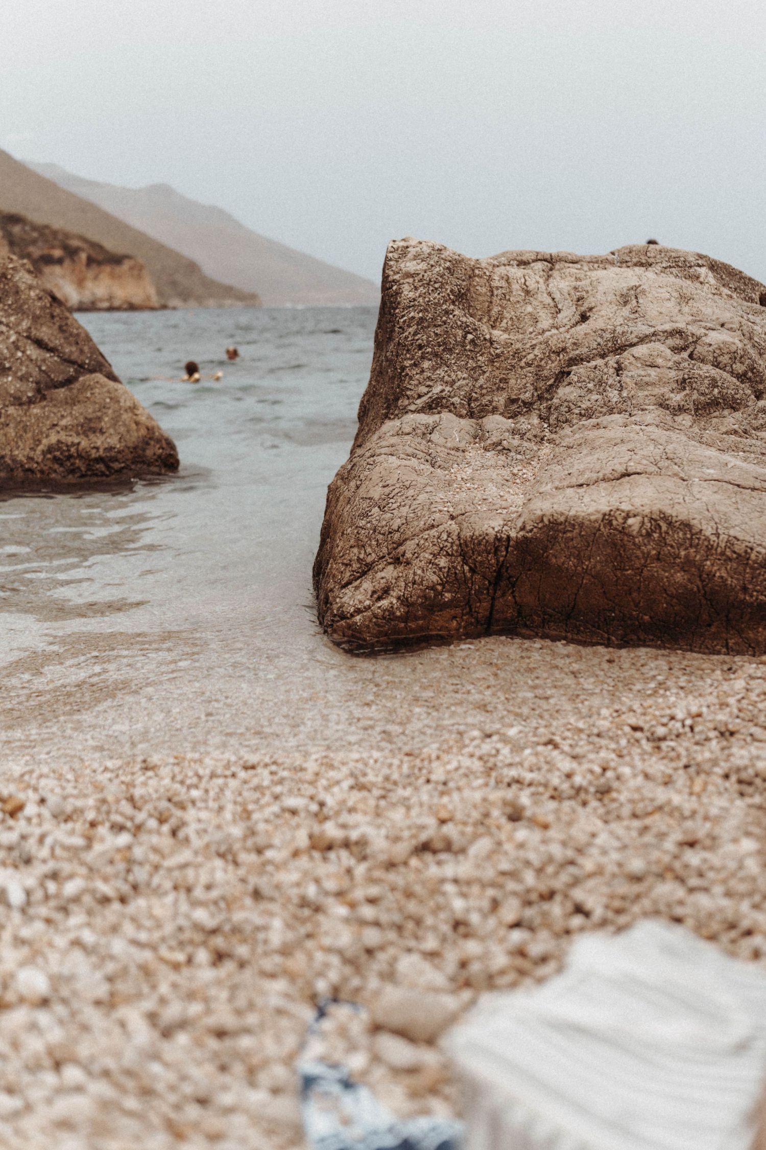 Large rocky boulders on a sandy beach with misty mountains and coastline in the background on an overcast day.
