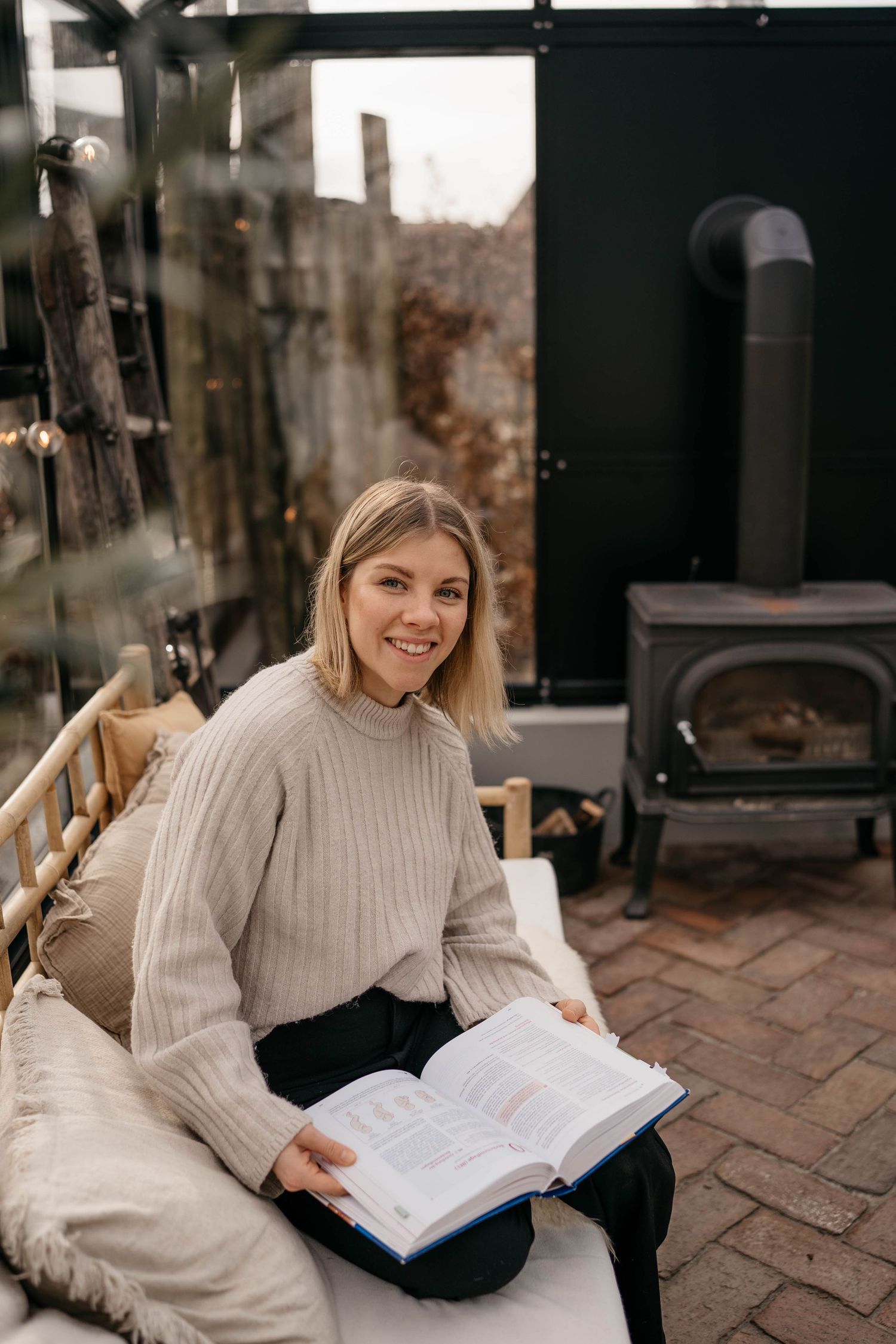 Figure in cozy sweater reads book while relaxing near wood stove on brick patio with plush seating.