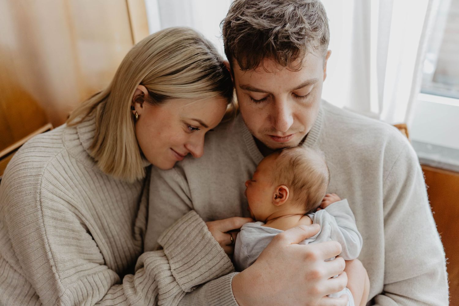 Intimate family moment with newborn baby in cozy sweaters by window light.