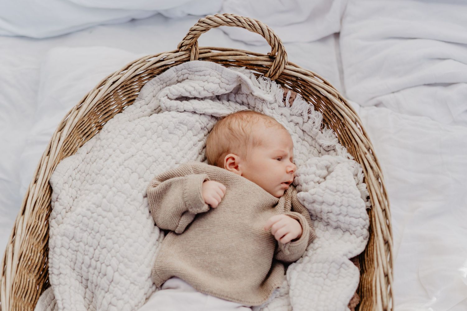 A sleeping infant wearing beige clothing lies peacefully in a woven basket on white textured blankets.