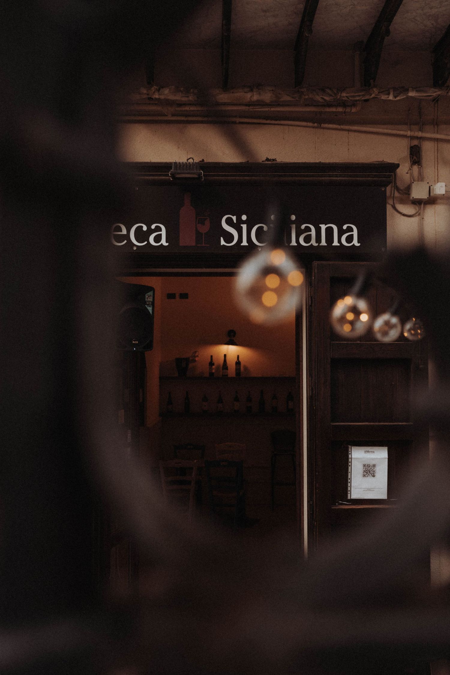 A dimly lit exterior view of a Sicilian cafe storefront with glowing pendant lights and partial signage visible through darkness.