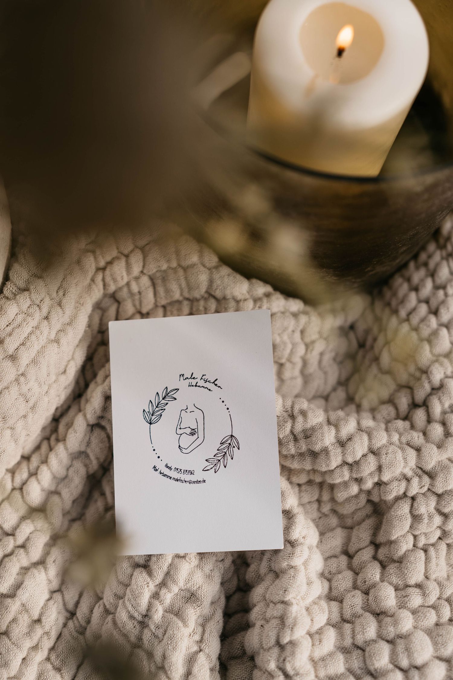 A white lit candle sits above a simple line drawing card on a textured knit blanket.
