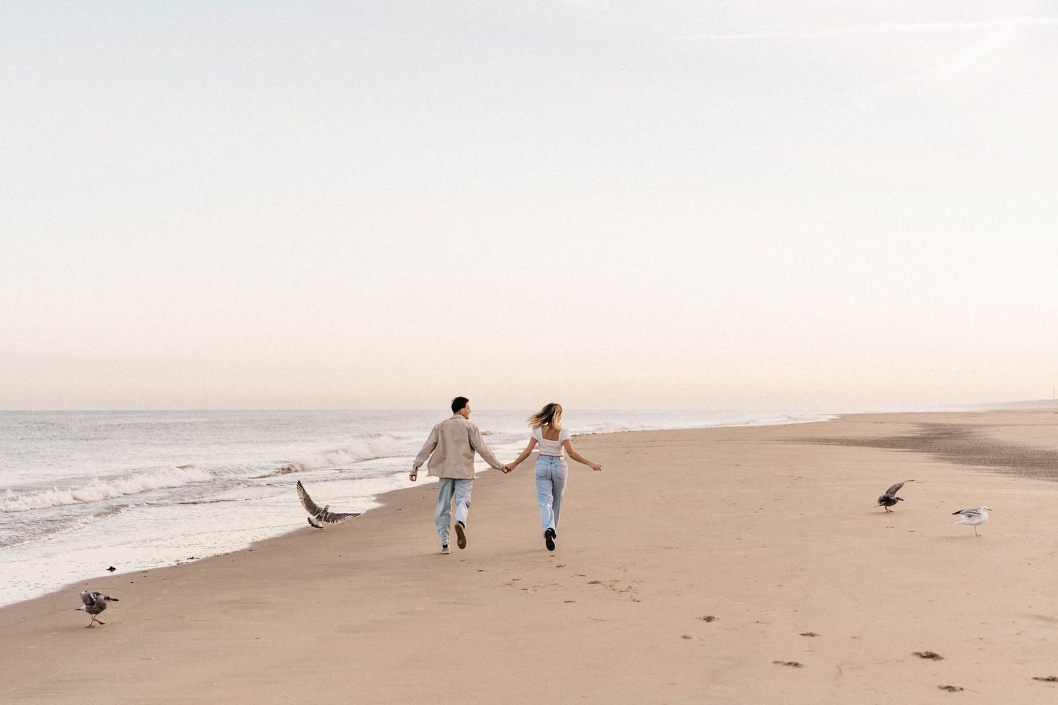 Couple running hand in hand along a peaceful beach at sunset with seagulls nearby.