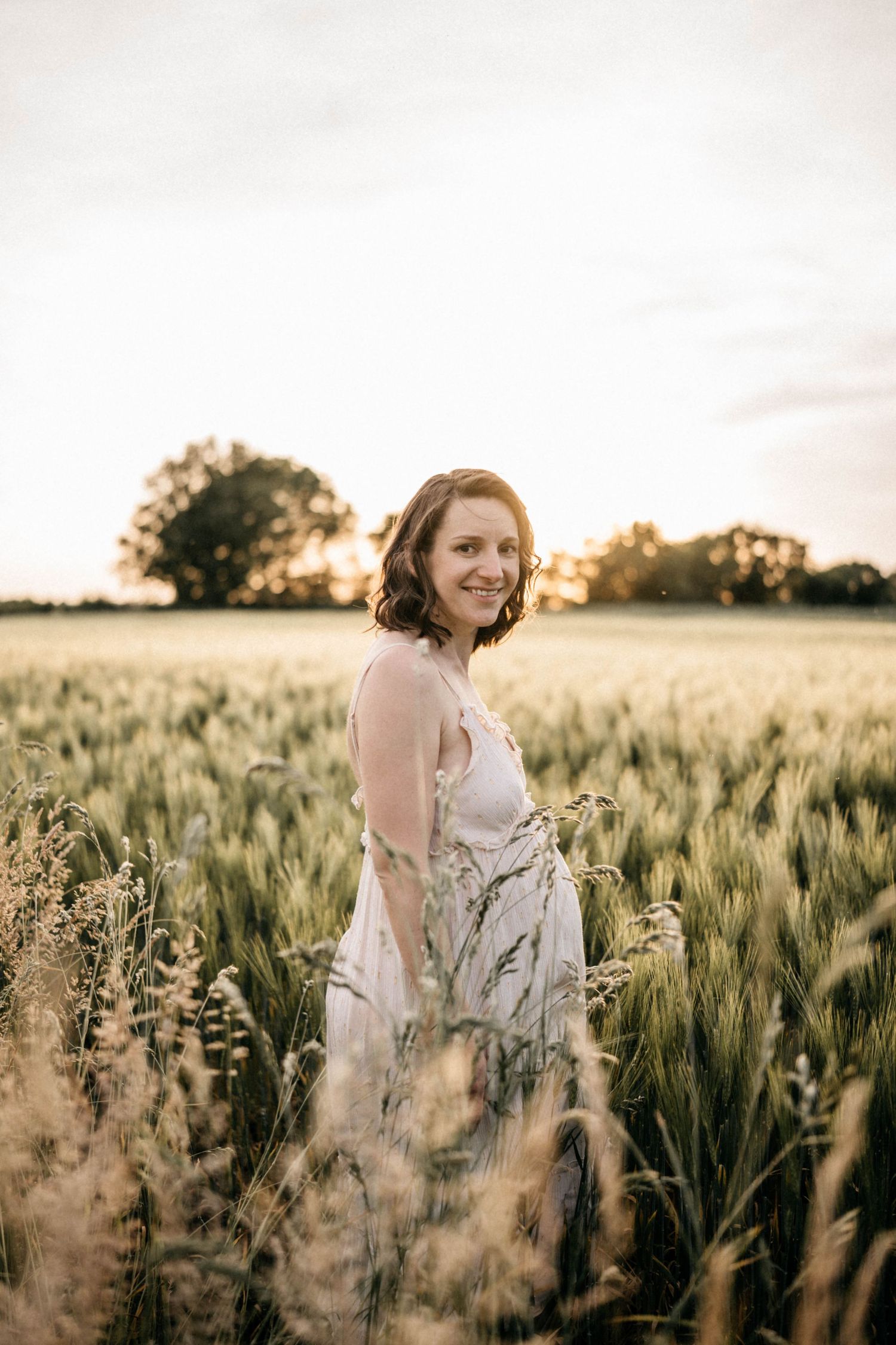 A woman in a white dress stands in a golden wheat field at sunset.