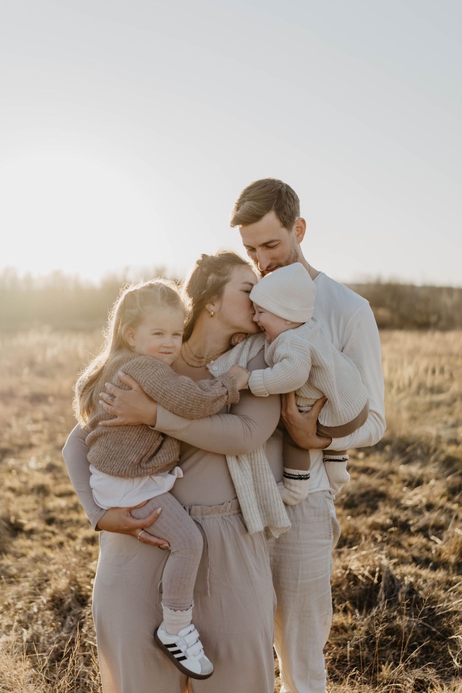 Family embracing in a sunlit field wearing neutral tones during golden hour.