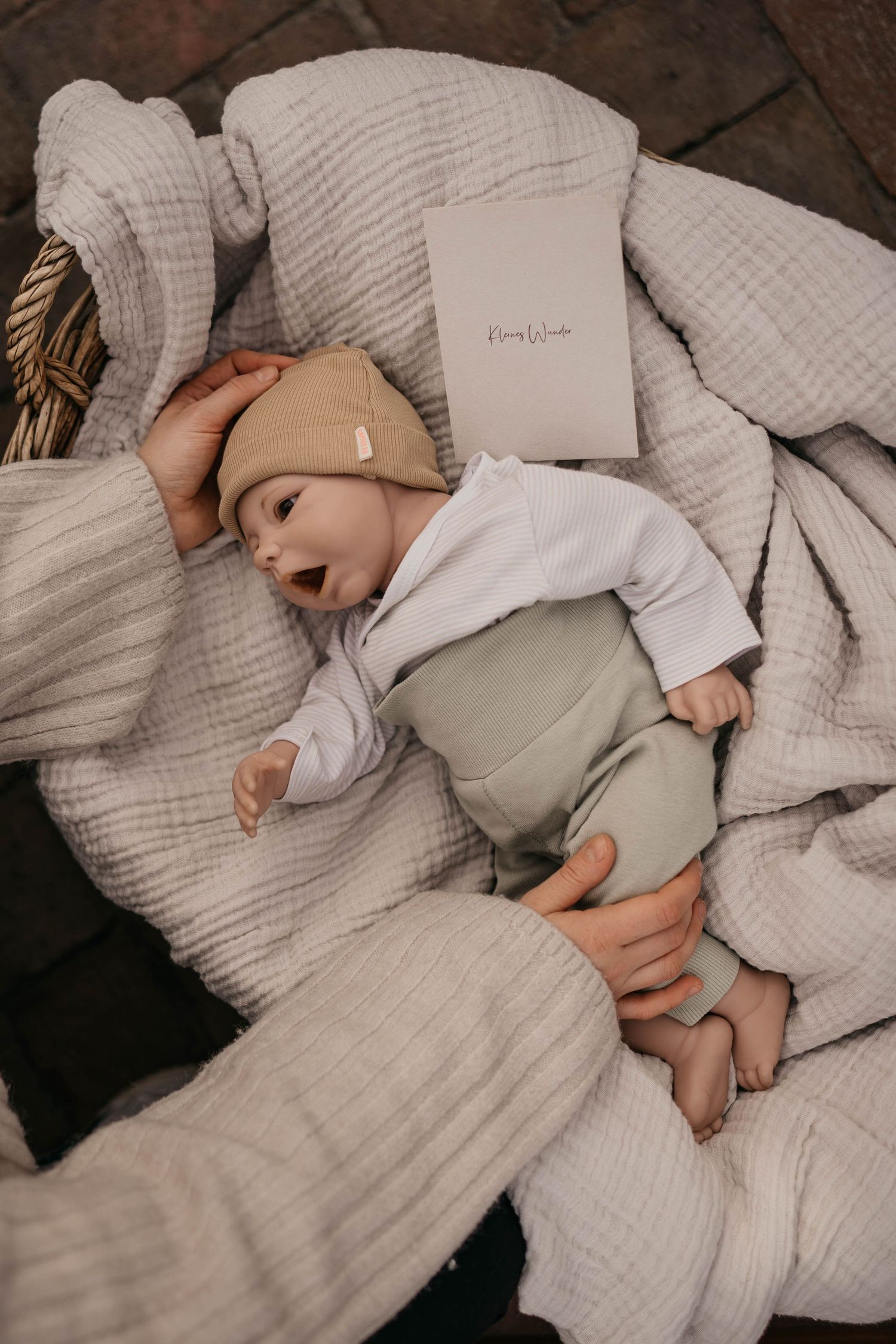 A baby in soft gray and white clothing lies on cozy textured bedding with neutral tones and natural textures.