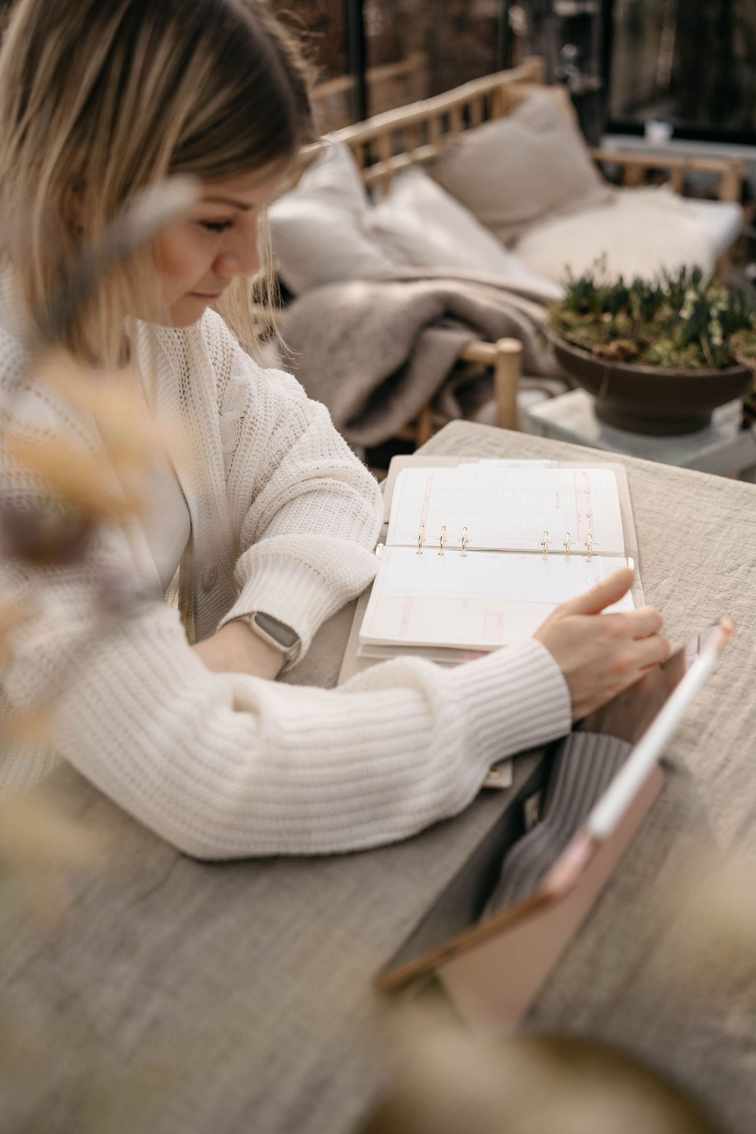 Someone in a cozy white sweater writes in a journal while sitting at a wooden table outdoors with cushions nearby.