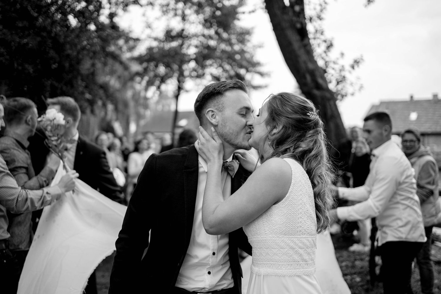 Black and white wedding photo showing newlyweds kissing during outdoor reception as guests dance in background.