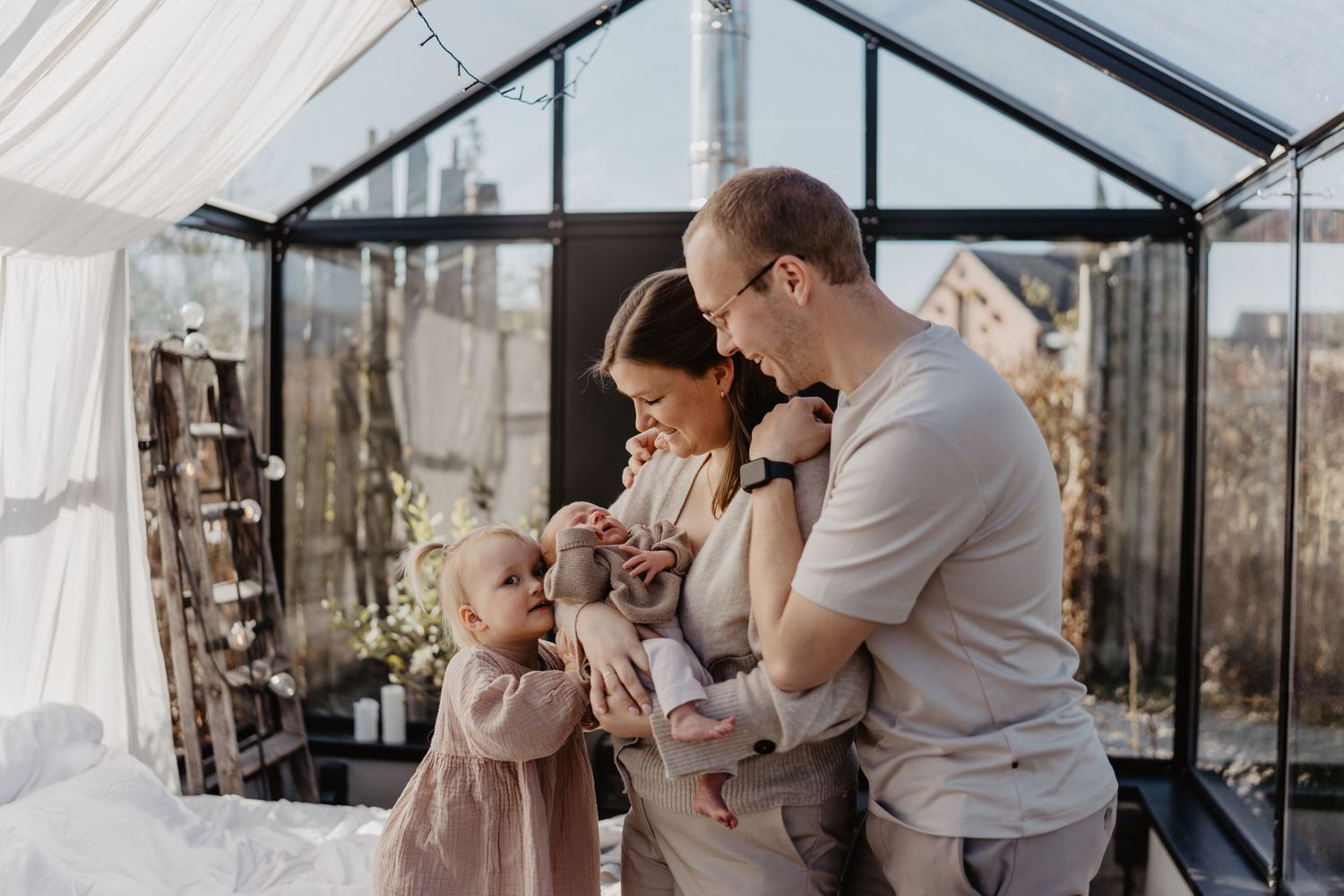 A family shares a tender moment together inside a greenhouse with natural light streaming through the glass panels.