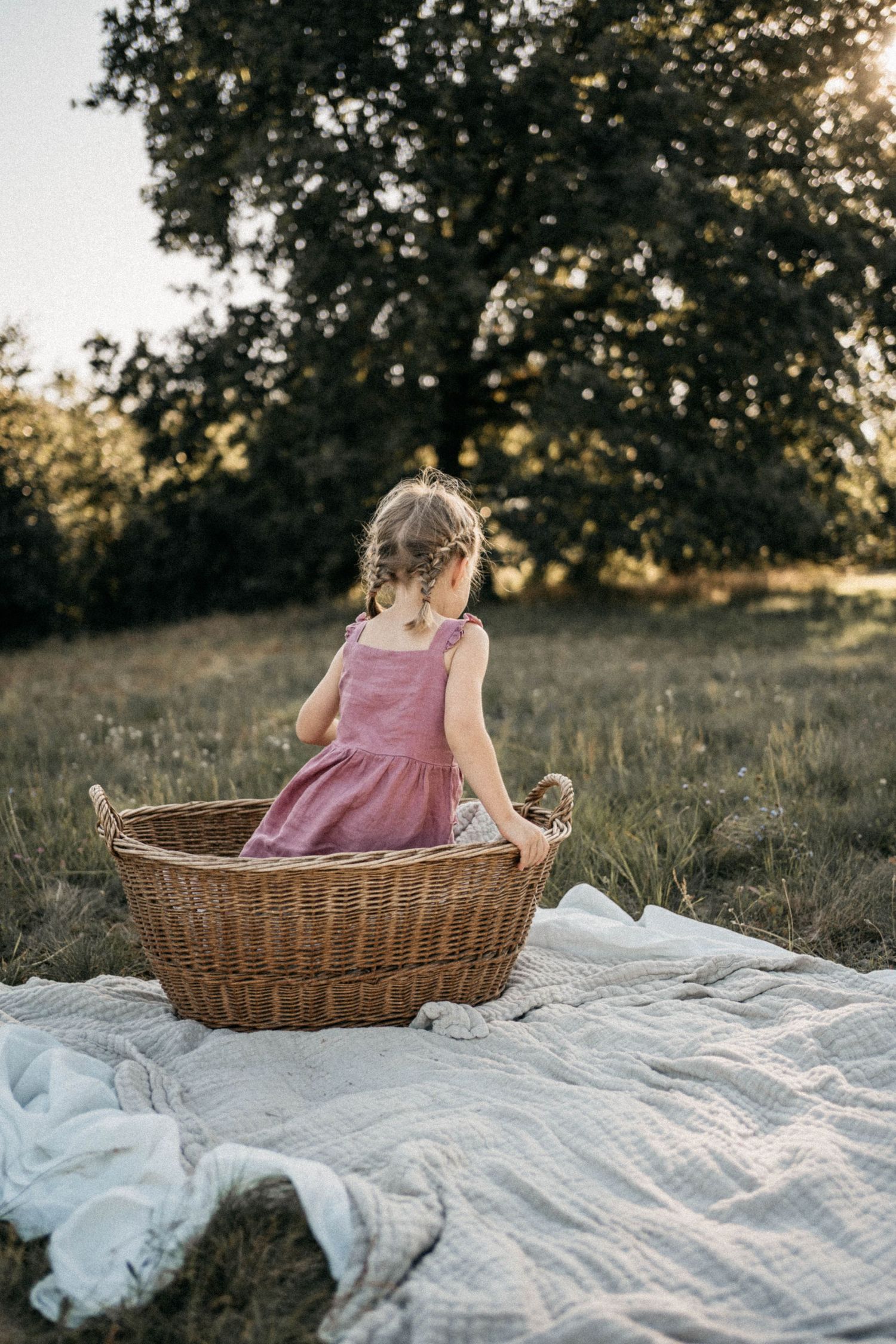 Child sitting in wicker basket on white blanket during sunset outdoors.