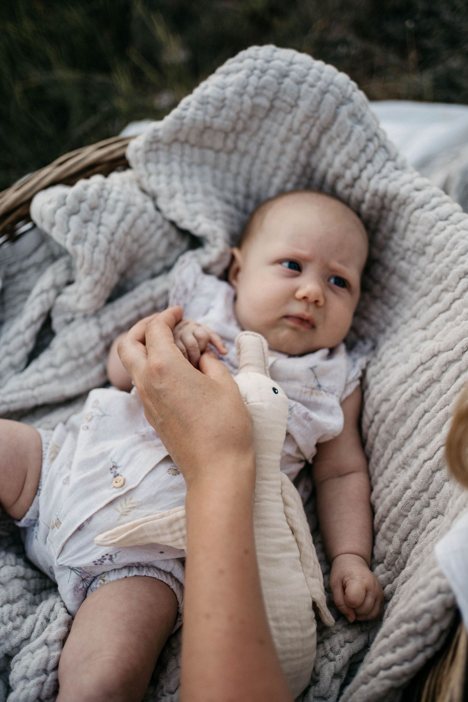 Baby lying on soft gray blanket in wicker basket outdoors.