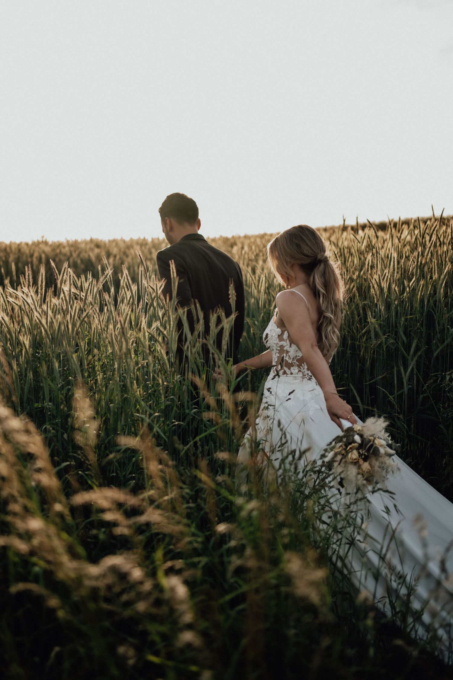 A couple walks through a golden wheat field at sunset, with the bride wearing a flowing white wedding dress.