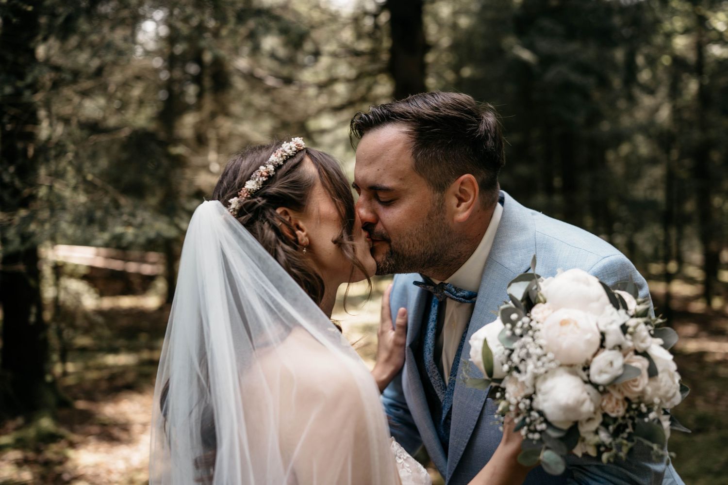 A romantic wedding moment captured in a sunlit forest with the couple sharing a tender kiss amidst nature.