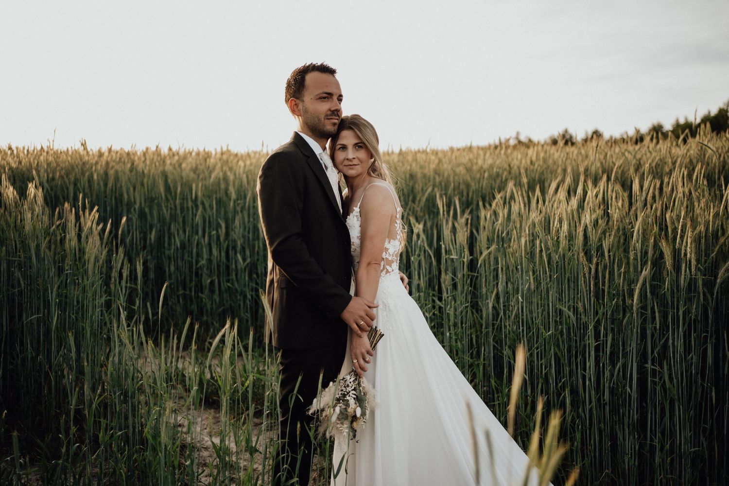 A romantic couple in wedding attire embrace while standing in a golden wheat field at sunset.