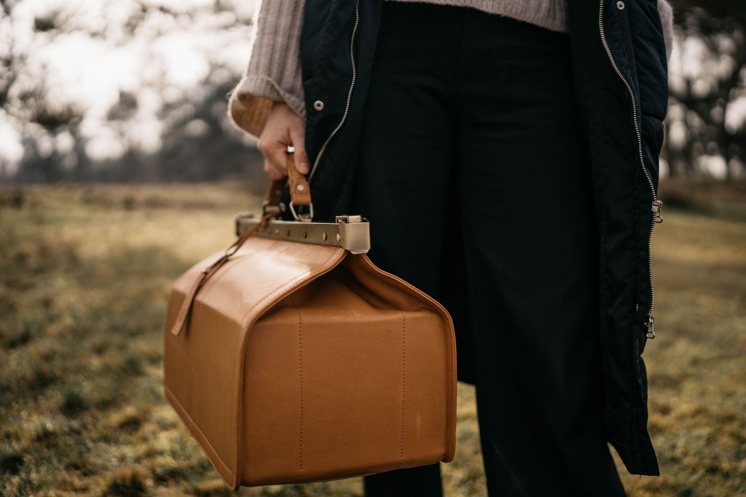Person holding a vintage brown leather doctor-style bag against a blurred natural outdoor background.