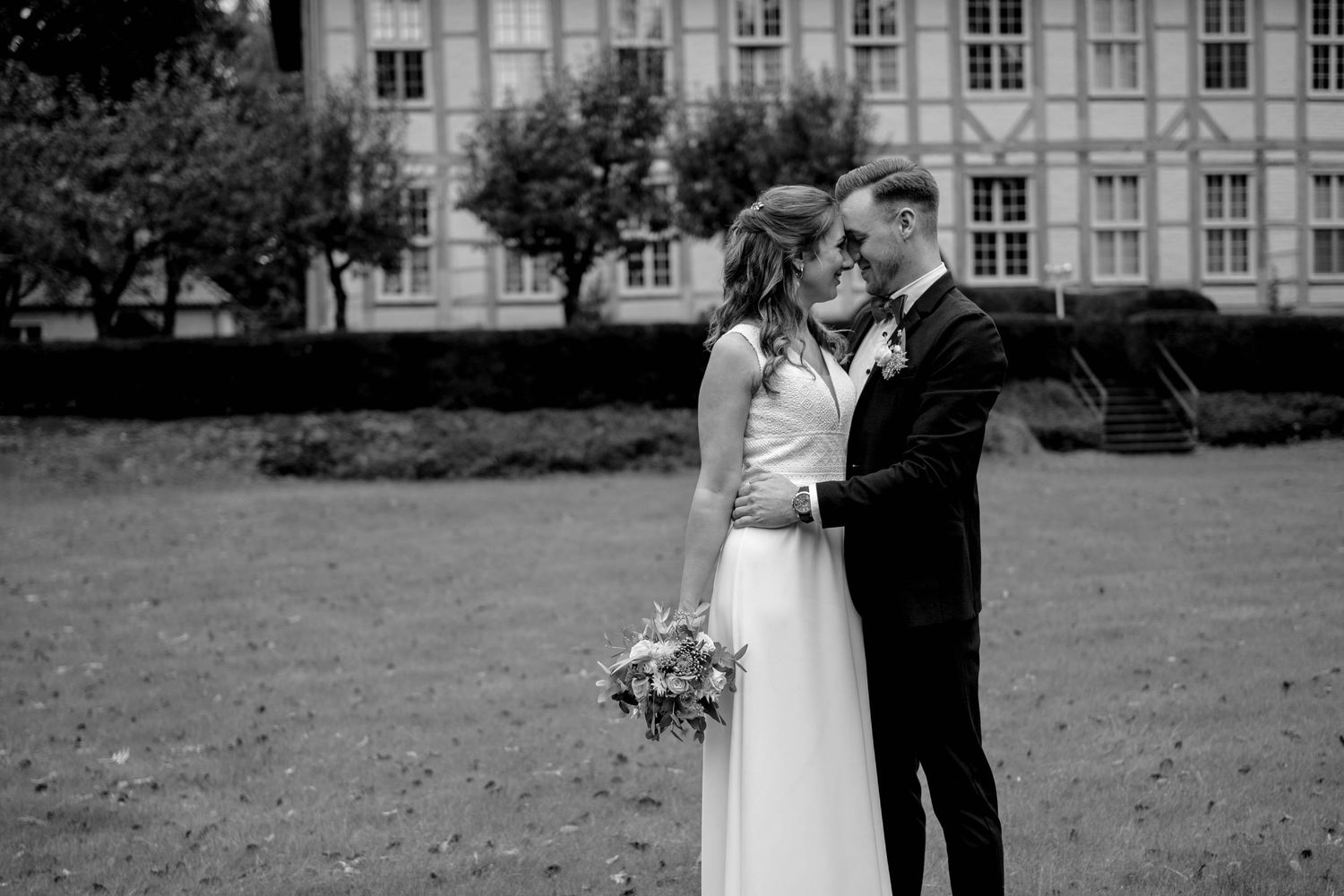 A couple in wedding attire share an intimate moment outdoors on the lawn in front of a grand building in black and white.