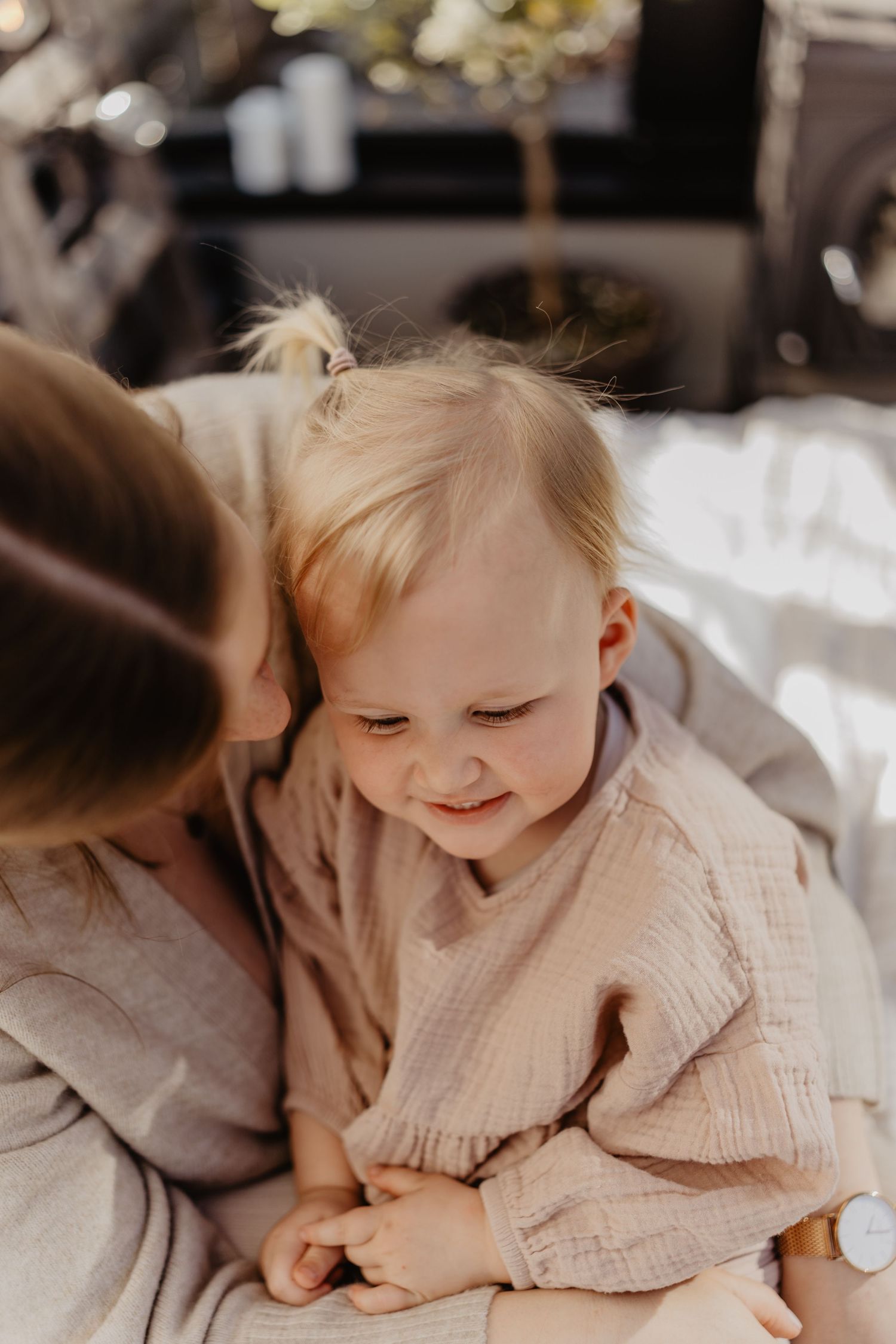 A baby in a light beige linen shirt smiles while sitting on a bed.