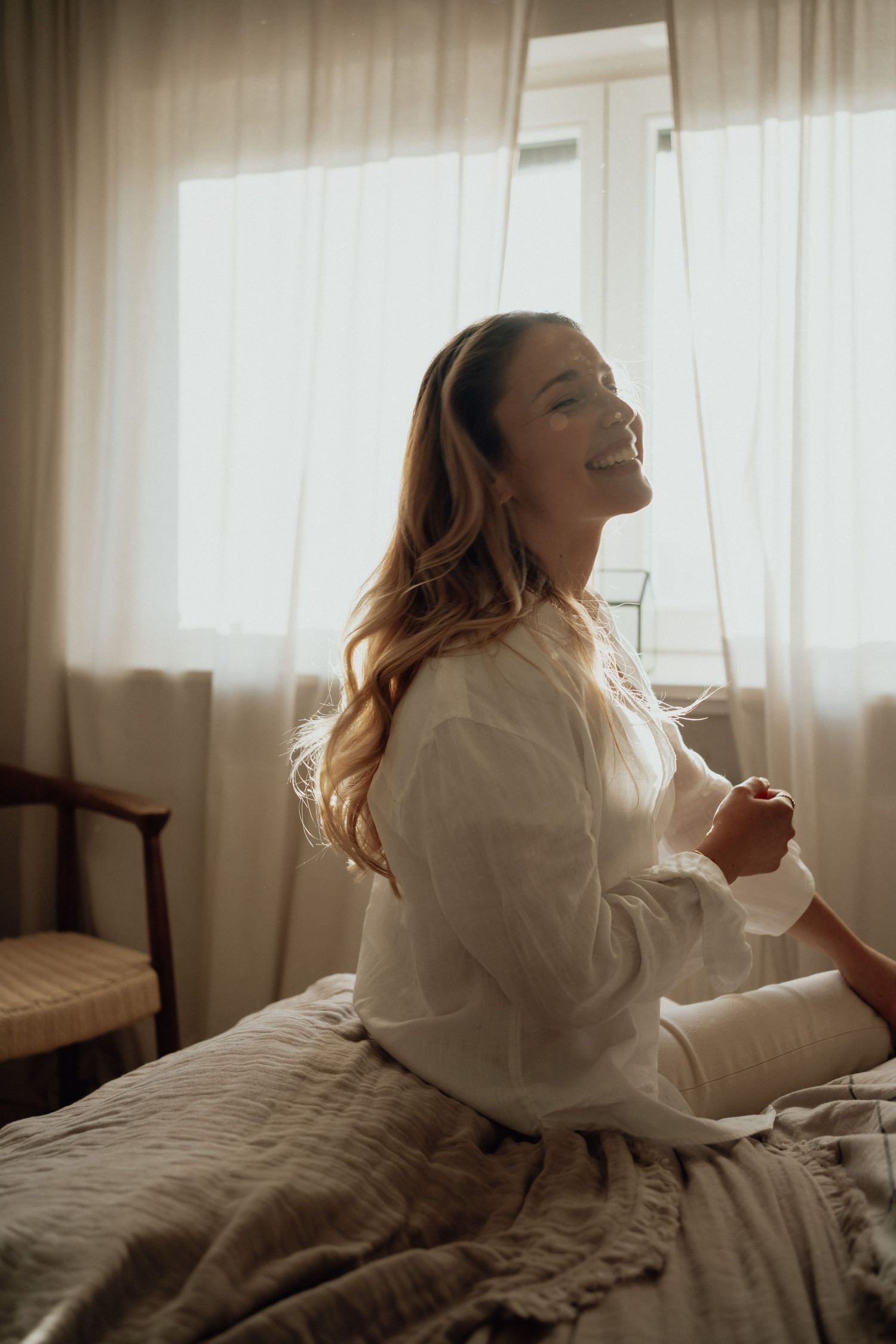 Woman in white sweater laughing joyfully while sitting on bed in sunlit room.
