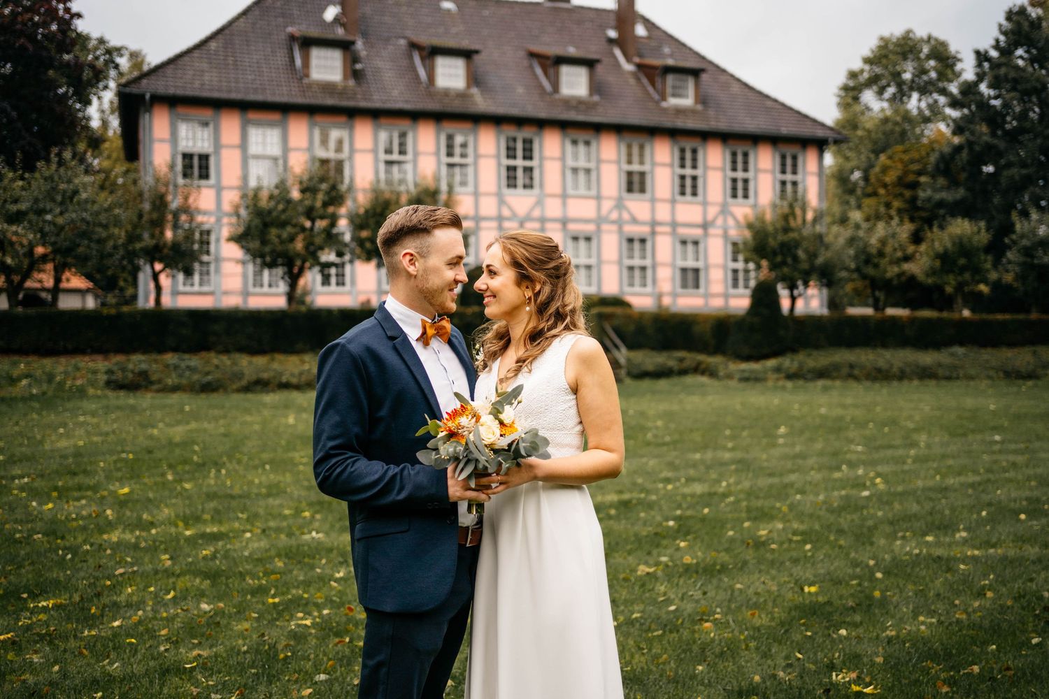 A couple shares a romantic moment while holding a bouquet in front of a historic pink mansion with a manicured lawn.