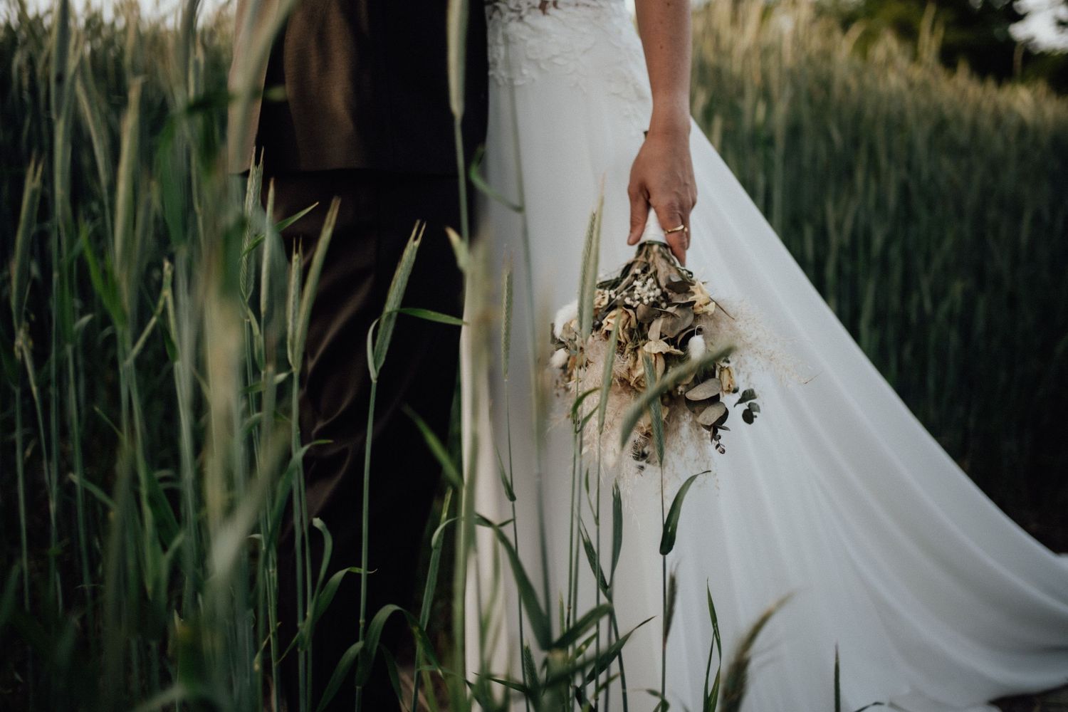 Elegant white bridal gown and bouquet amid tall green grass at sunset in rustic outdoor wedding setting.
