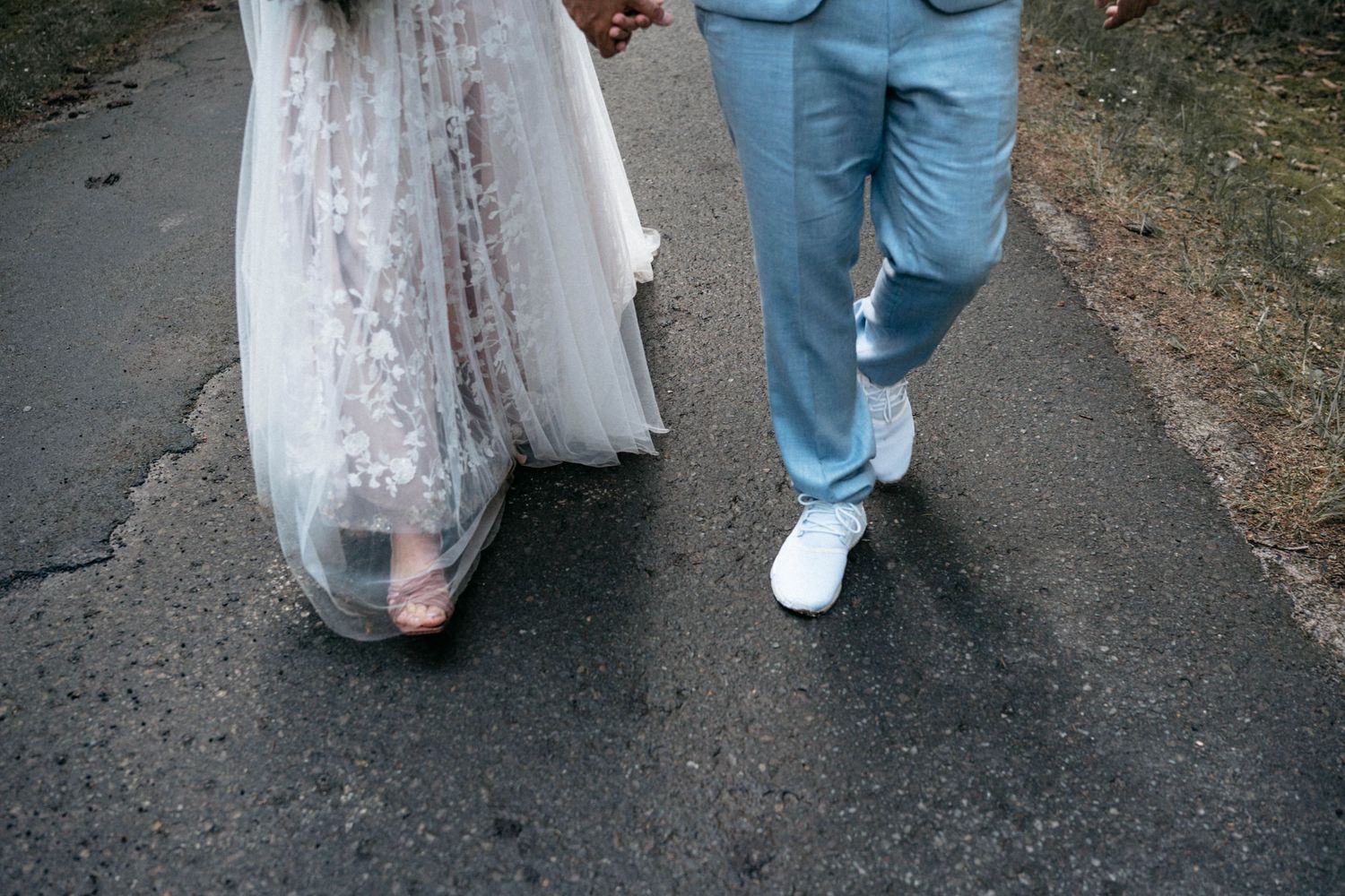A close-up view of newlyweds walking on a path, showing the white lace wedding dress hem and groom's light blue pants.