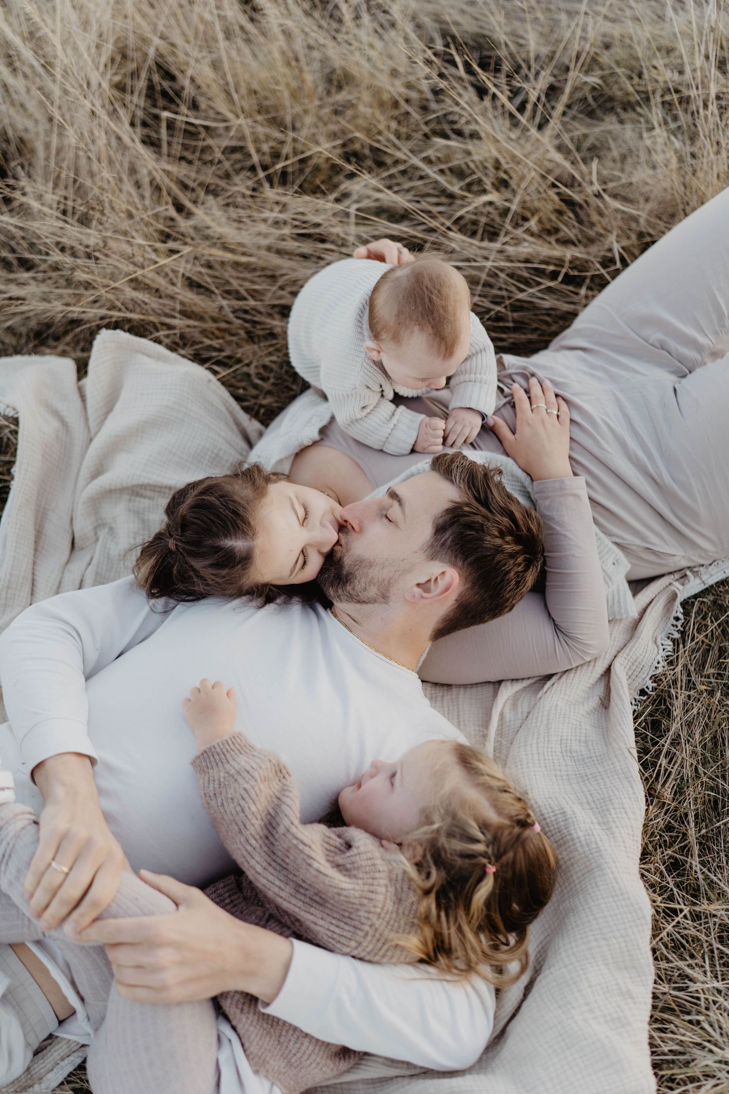 Family in cozy neutral colored sweaters lying together on a blanket in dried grass during an outdoor photo session.