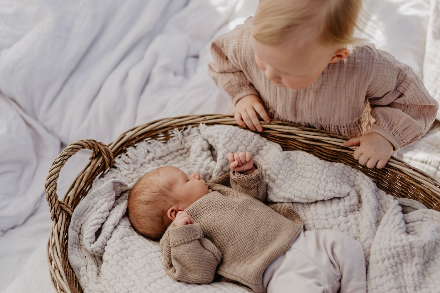 A young child peers into a woven basket containing a sleeping baby swaddled in neutral-colored blankets.