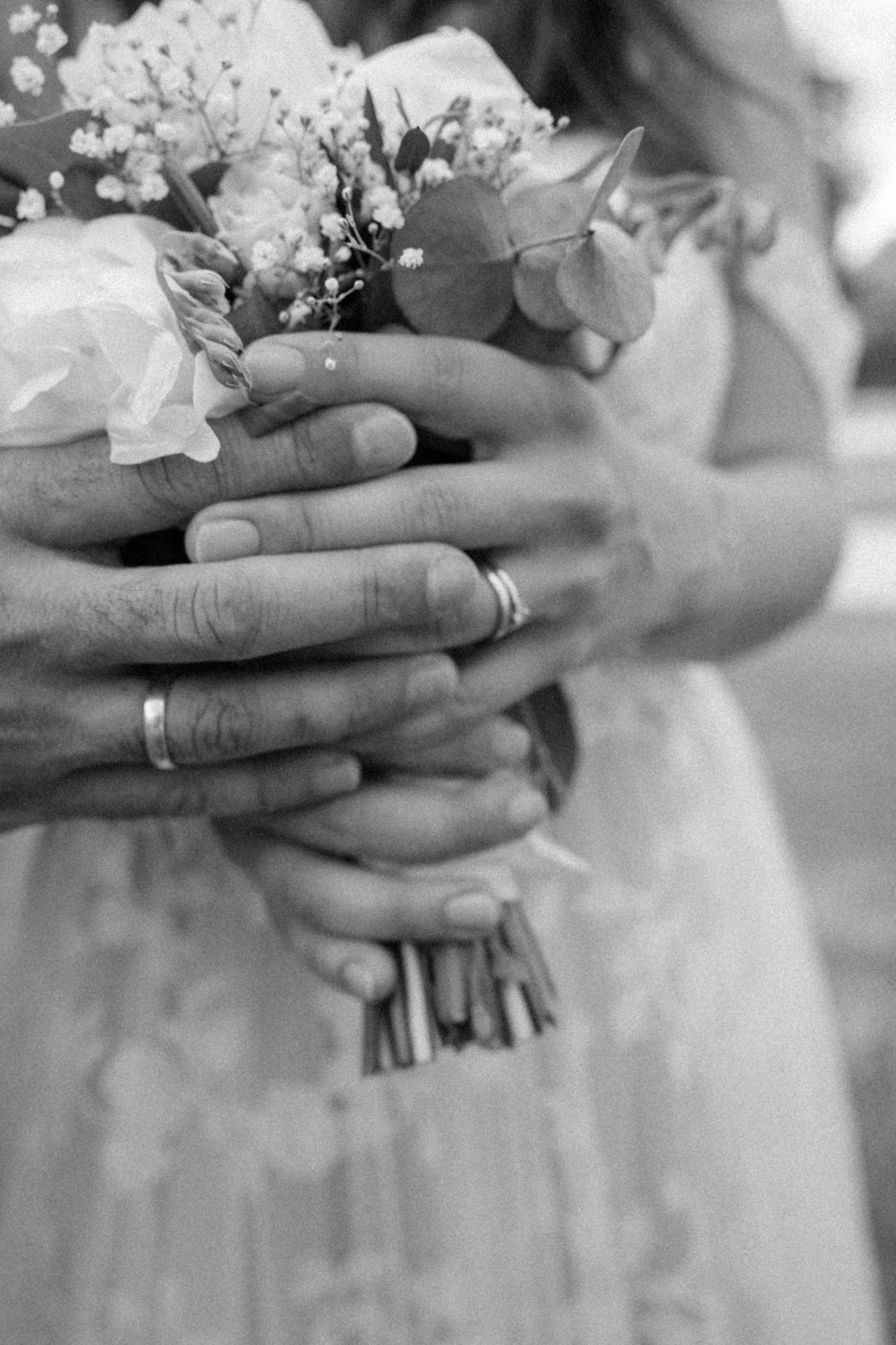 A close-up black and white shot of hands holding a delicate wedding bouquet with rings visible on the fingers.