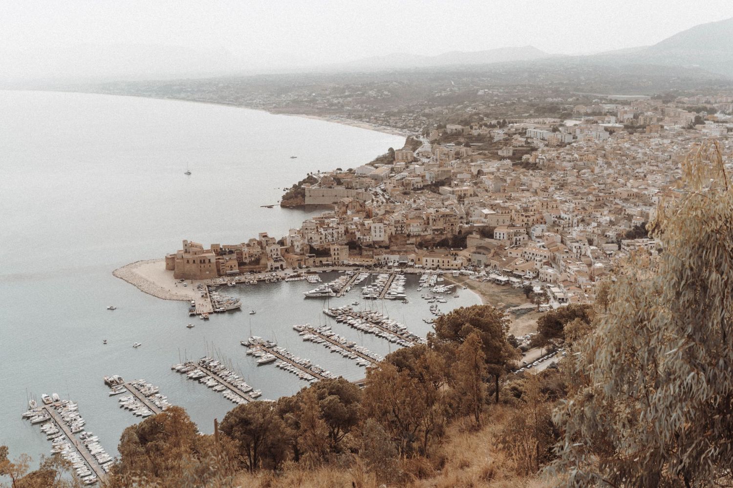 Aerial view of coastal Mediterranean marina with boats docked in harbor and hillside town overlooking the bay.
