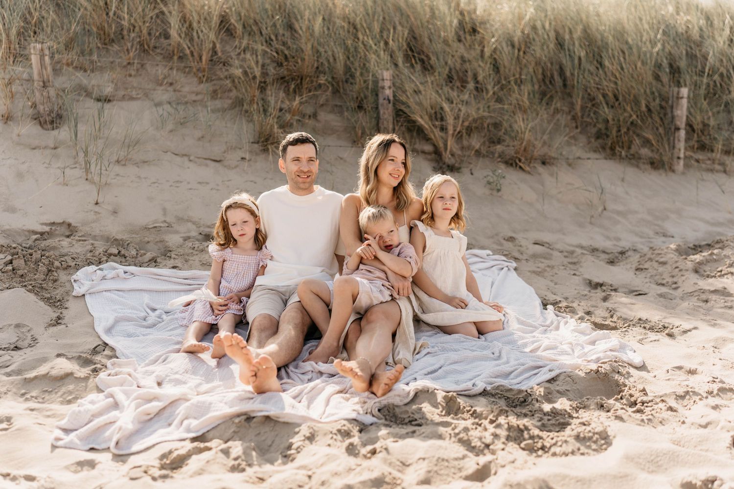 Family sitting together on a white blanket at the beach near sand dunes.