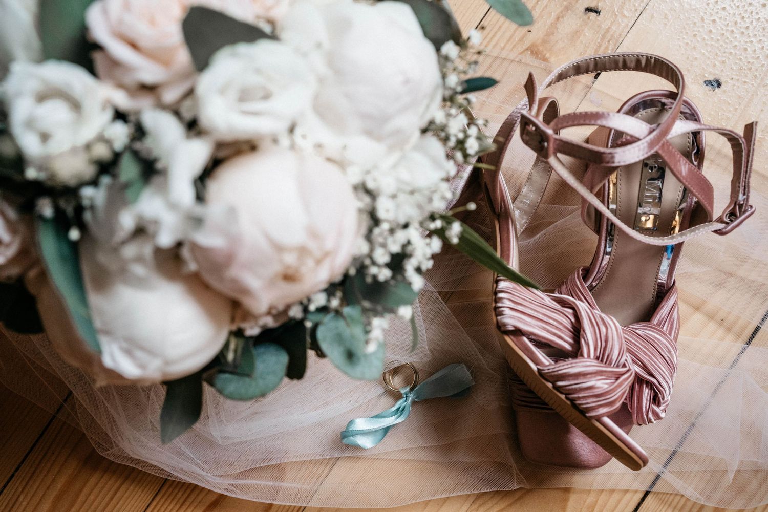 Elegant pink satin bridal shoes next to soft white peony bouquet on wooden floor.