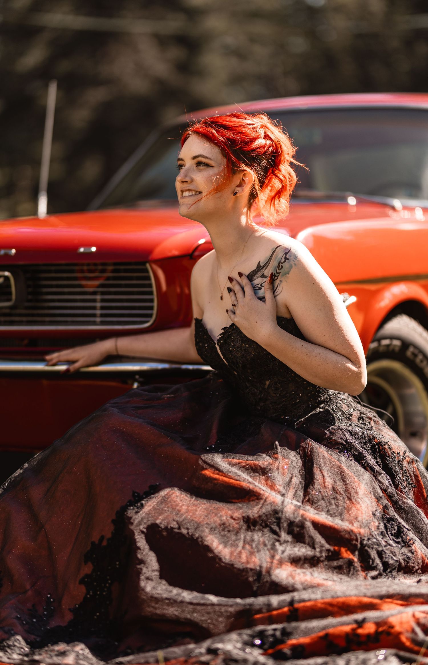 Red-haired woman in black ballgown poses beside a classic red Mustang, smiling outdoors.