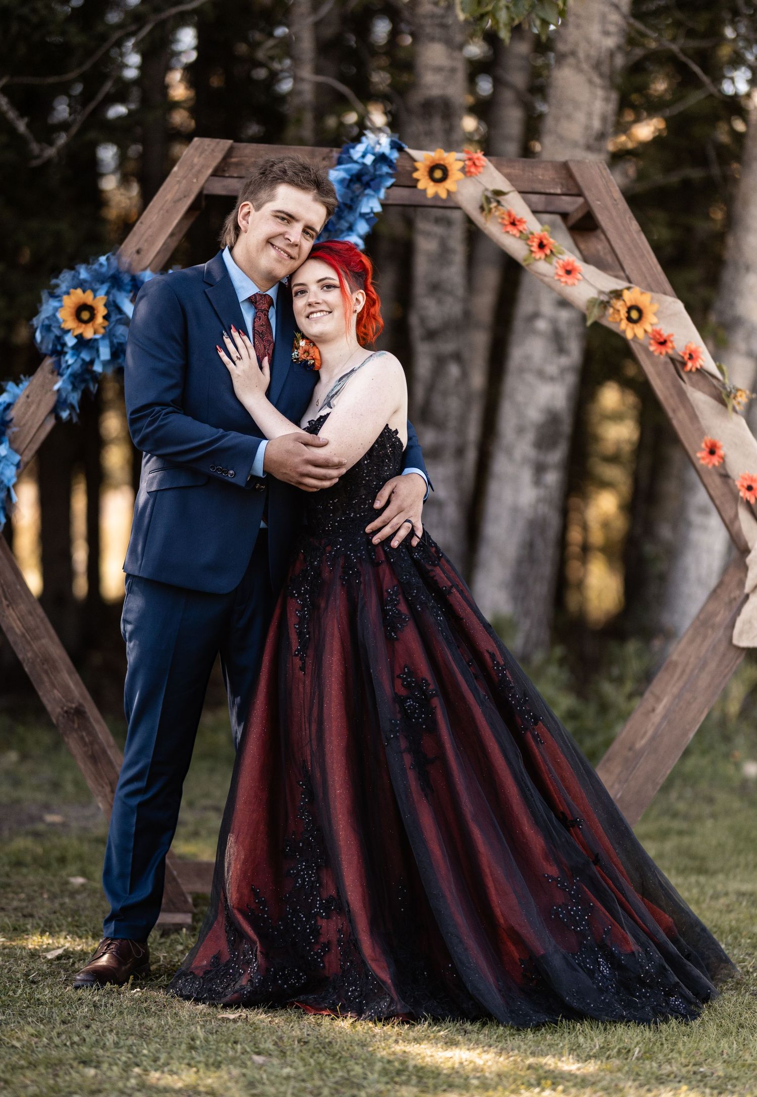 Couple posing before hexagonal wooden arch with sunflowers, bride in black-red gown, groom in navy suit.