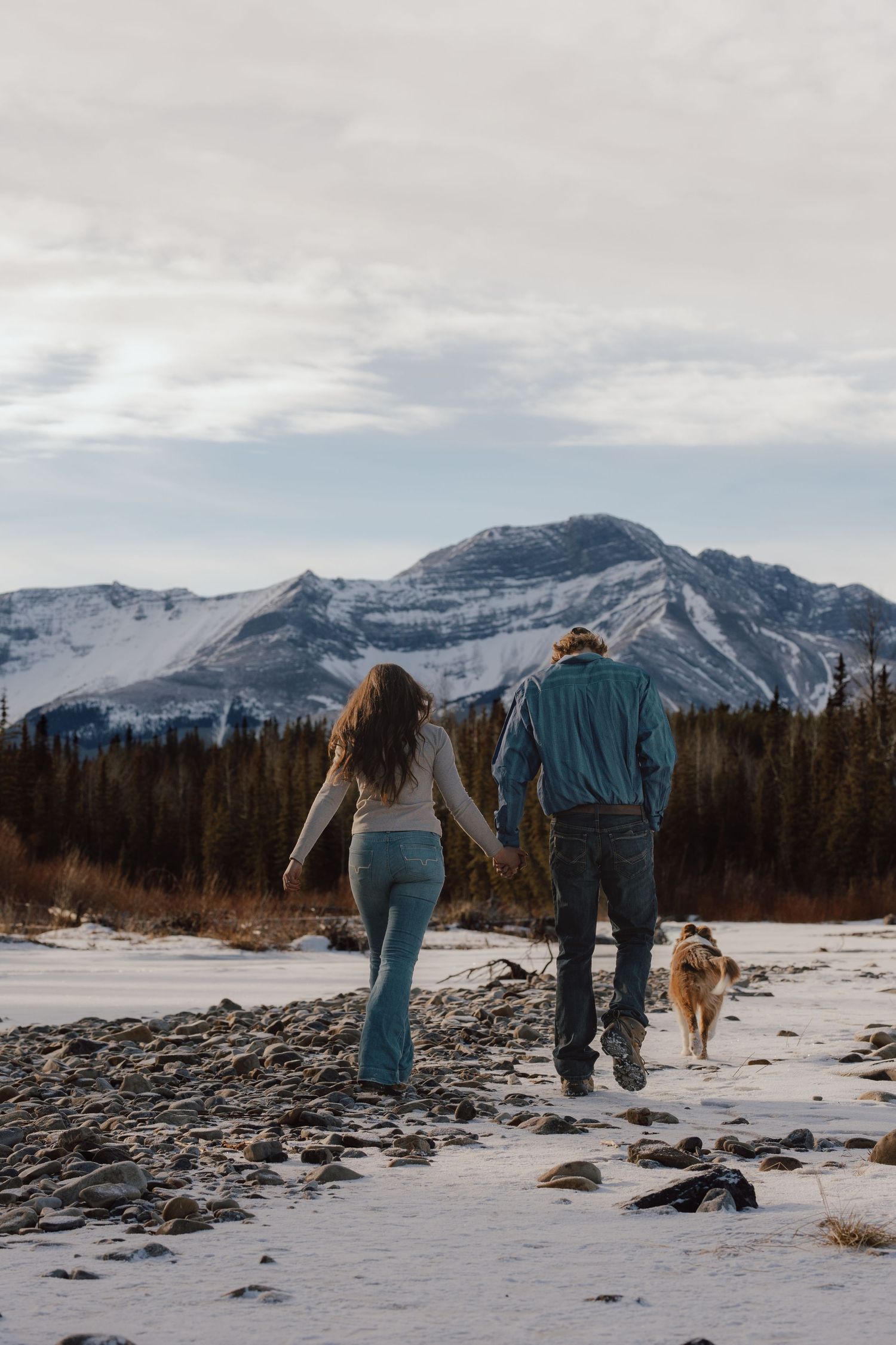 Couple holding hands walking with dog in snowy mountain landscape with evergreen trees and cloudy sky.