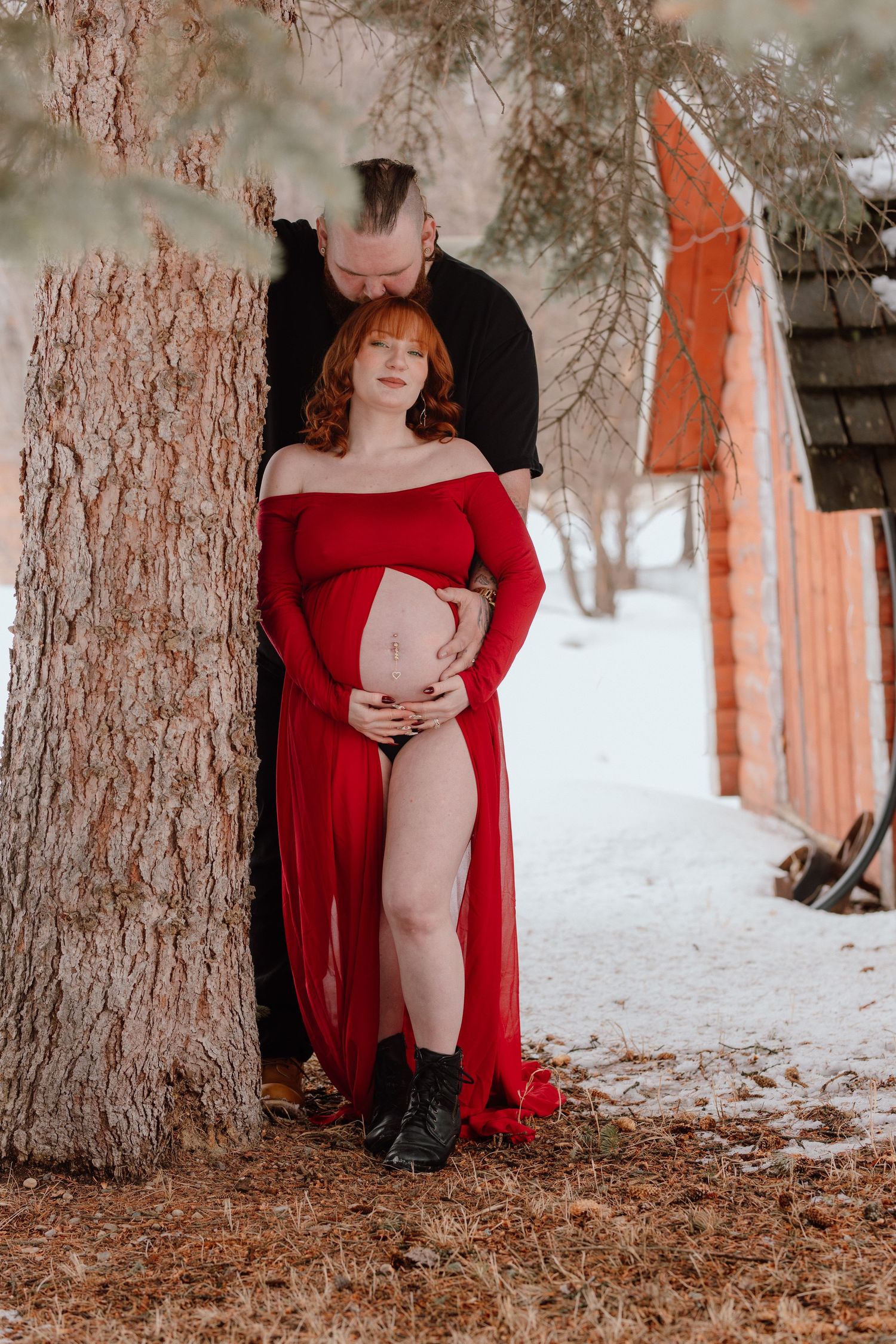 Couple posing by a tree in winter, pregnant woman in red dress cradling her baby bump with partner kissing her head.