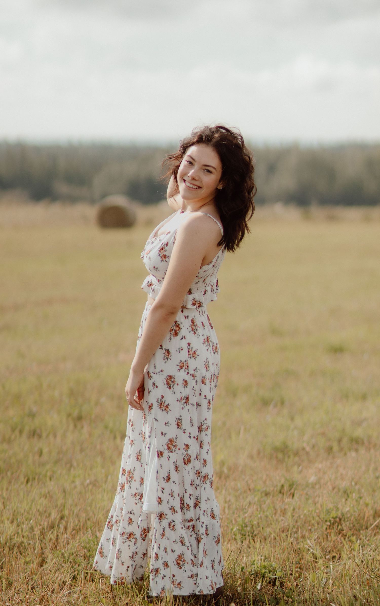 Smiling brunette woman in white floral maxi dress standing in an open field with hay bales in the background.