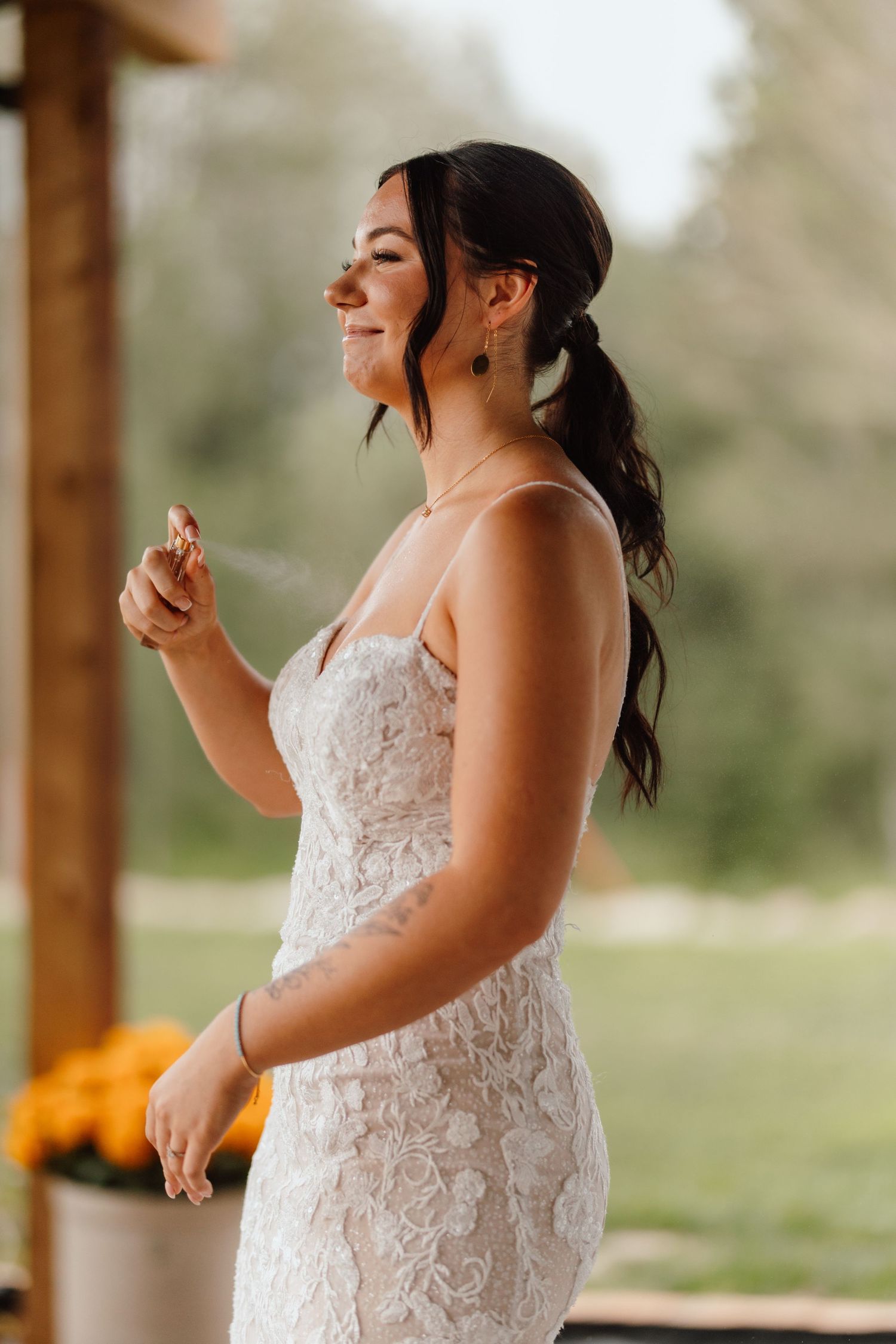 Smiling bride in lace wedding gown standing outdoors on a porch, spraying perfume on herself with flowers in background.