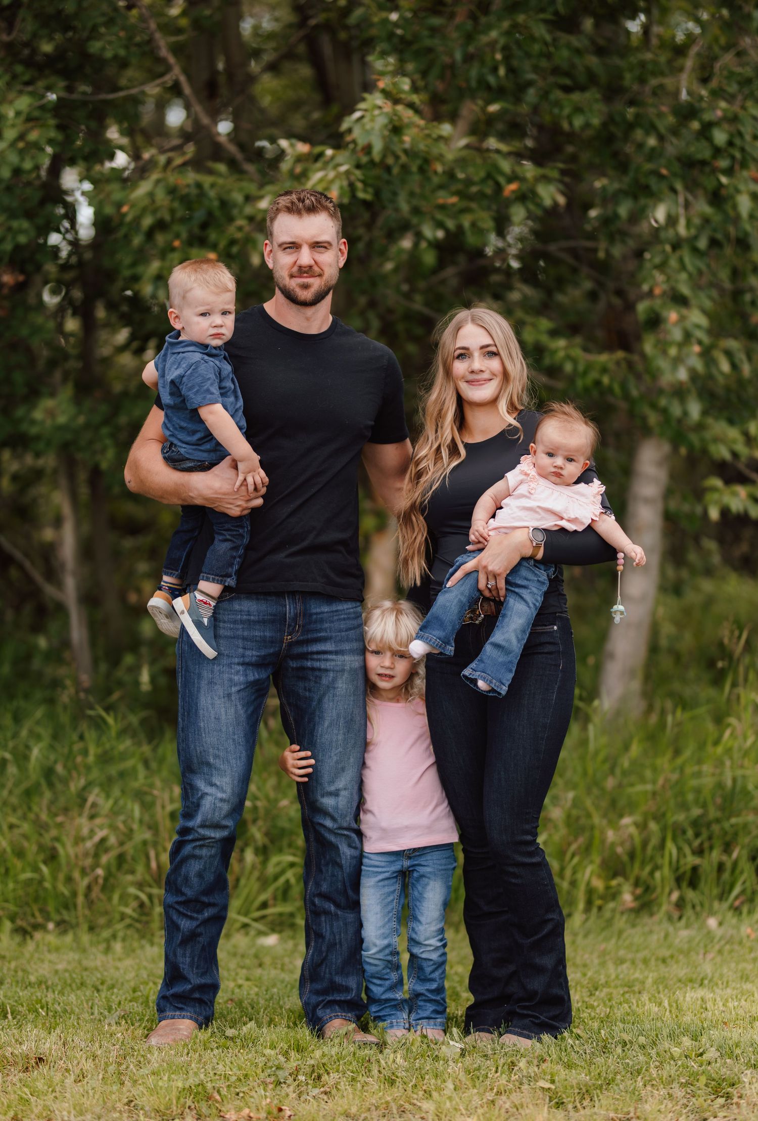 Family of five posing outdoors with two adults holding babies and a young girl standing between them.